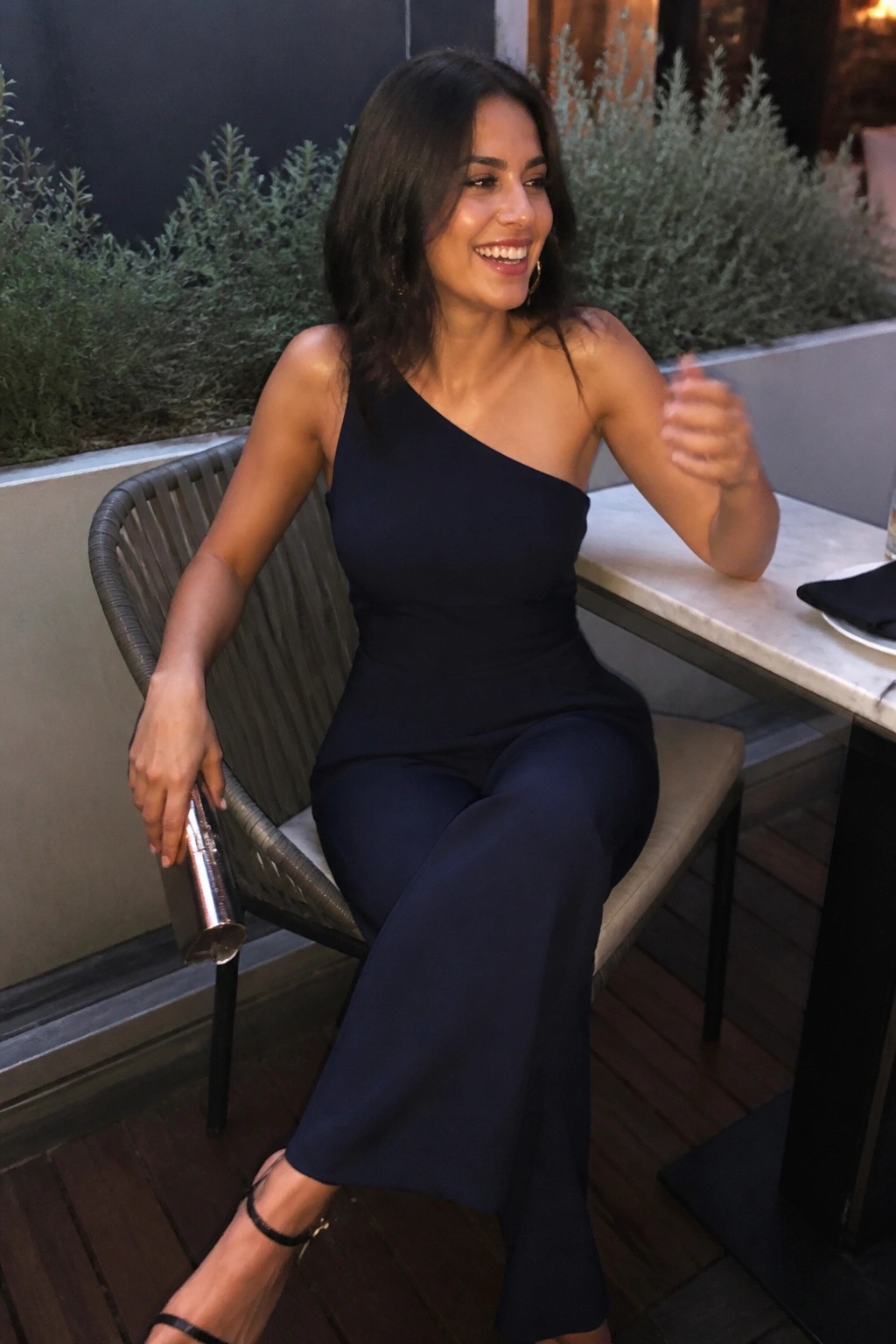 Smiling woman in navy one-shoulder jumpsuit seated outdoors holding metallic clutch.