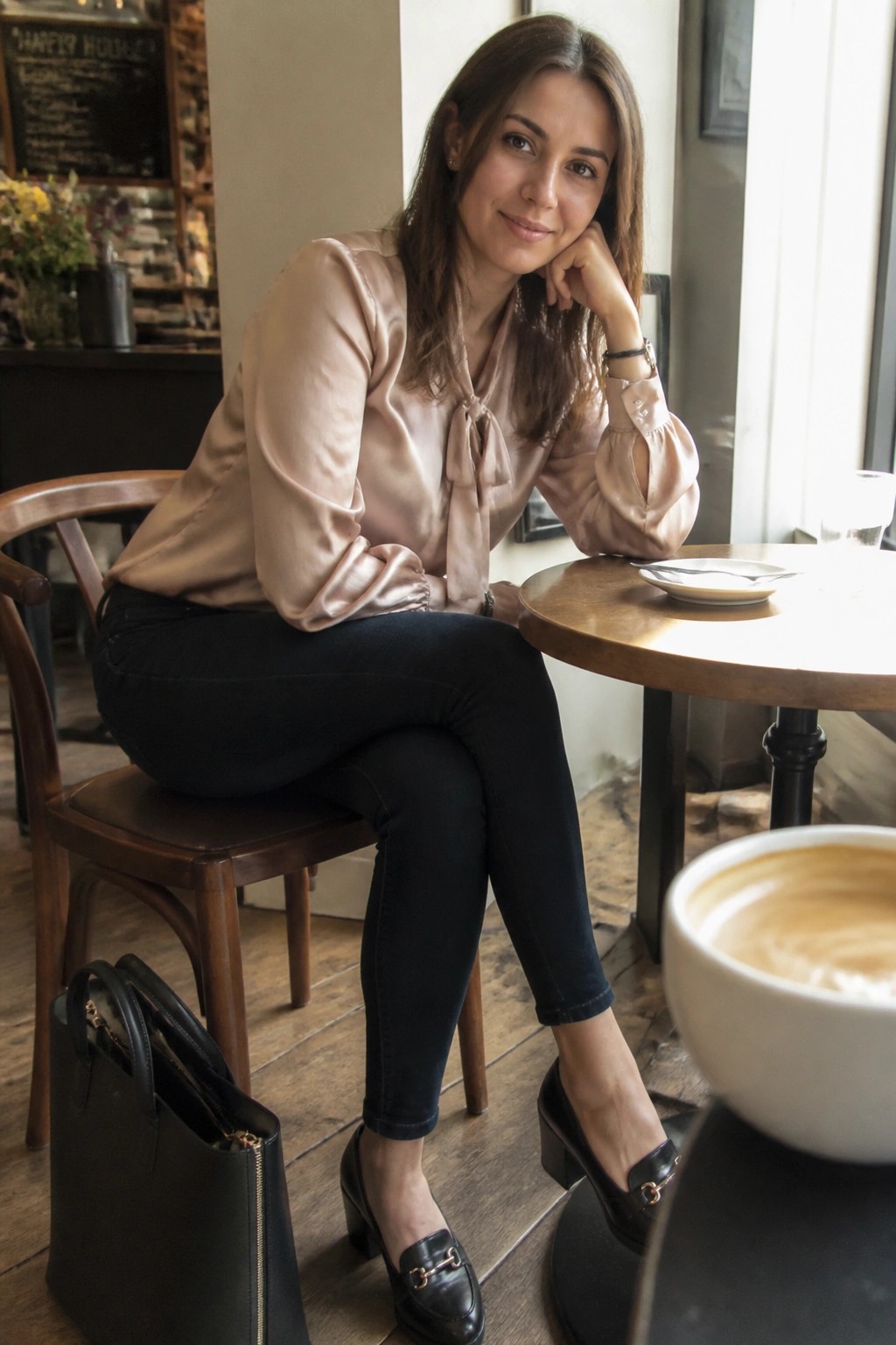 Woman in light pink satin blouse seated at cafe table with coffee and handbag