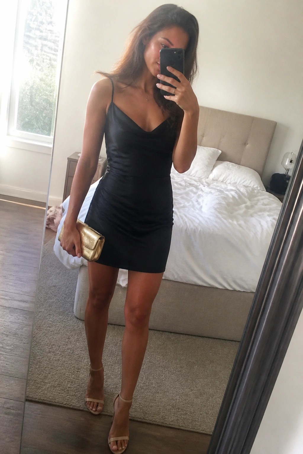 Woman in black mini dress taking mirror selfie in bedroom with gold clutch
