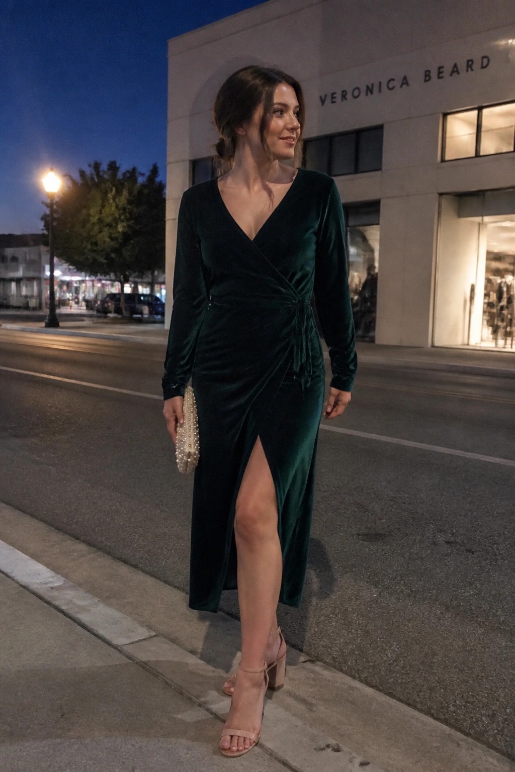 Woman in dark green velvet wrap dress holding clutch on nighttime sidewalk
