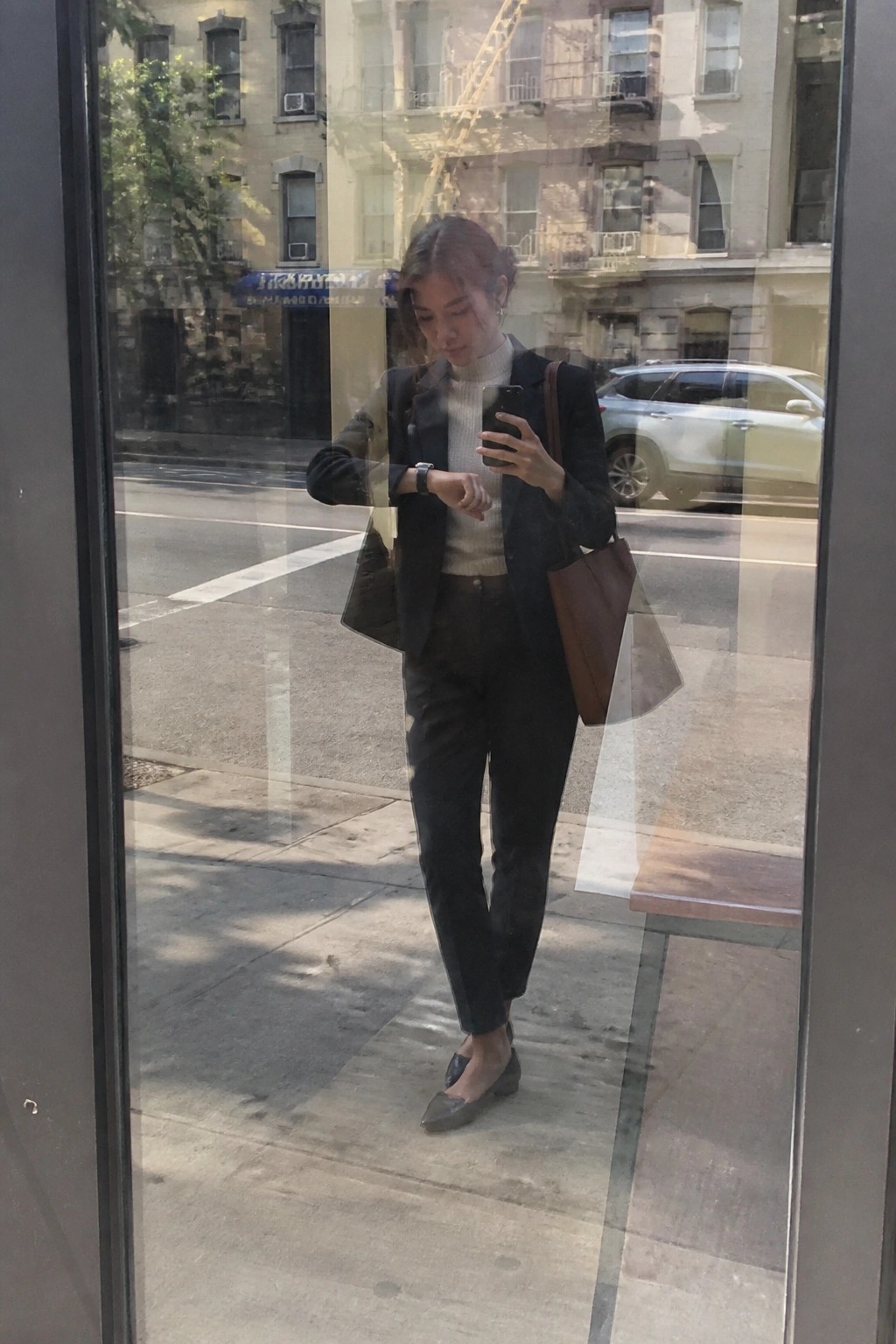 Woman in black blazer and turtleneck checks phone on city street reflection.