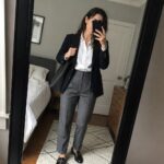 Woman in navy blazer and gray trousers taking mirror selfie in bedroom.