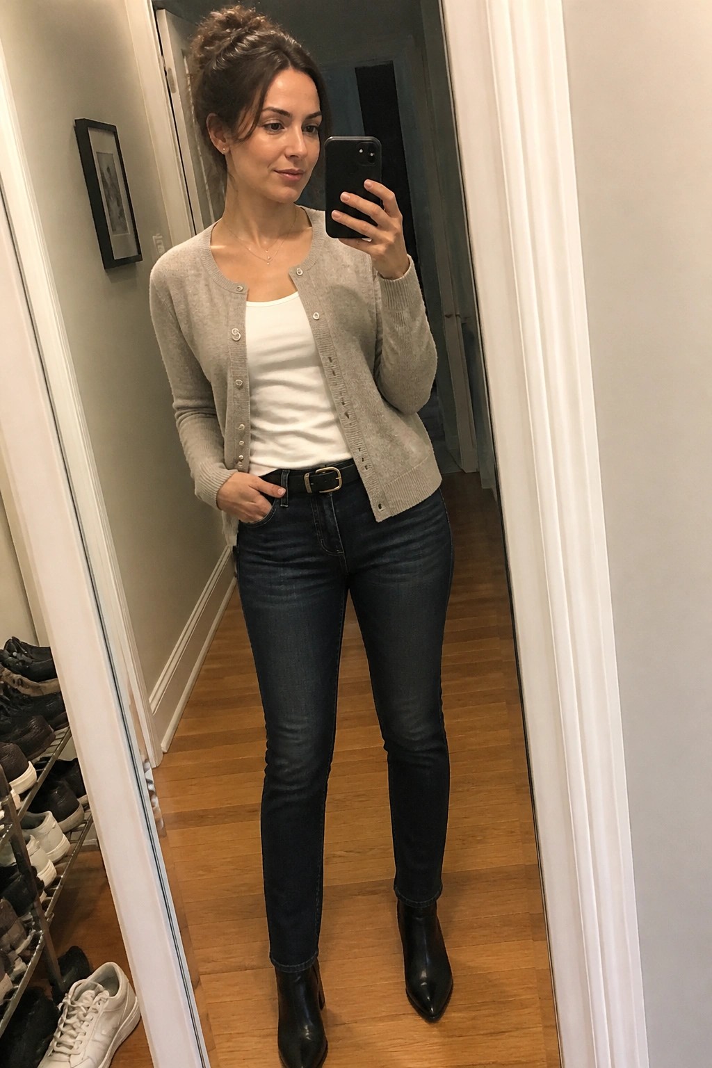 Woman in gray cardigan and dark jeans taking mirror selfie in hallway