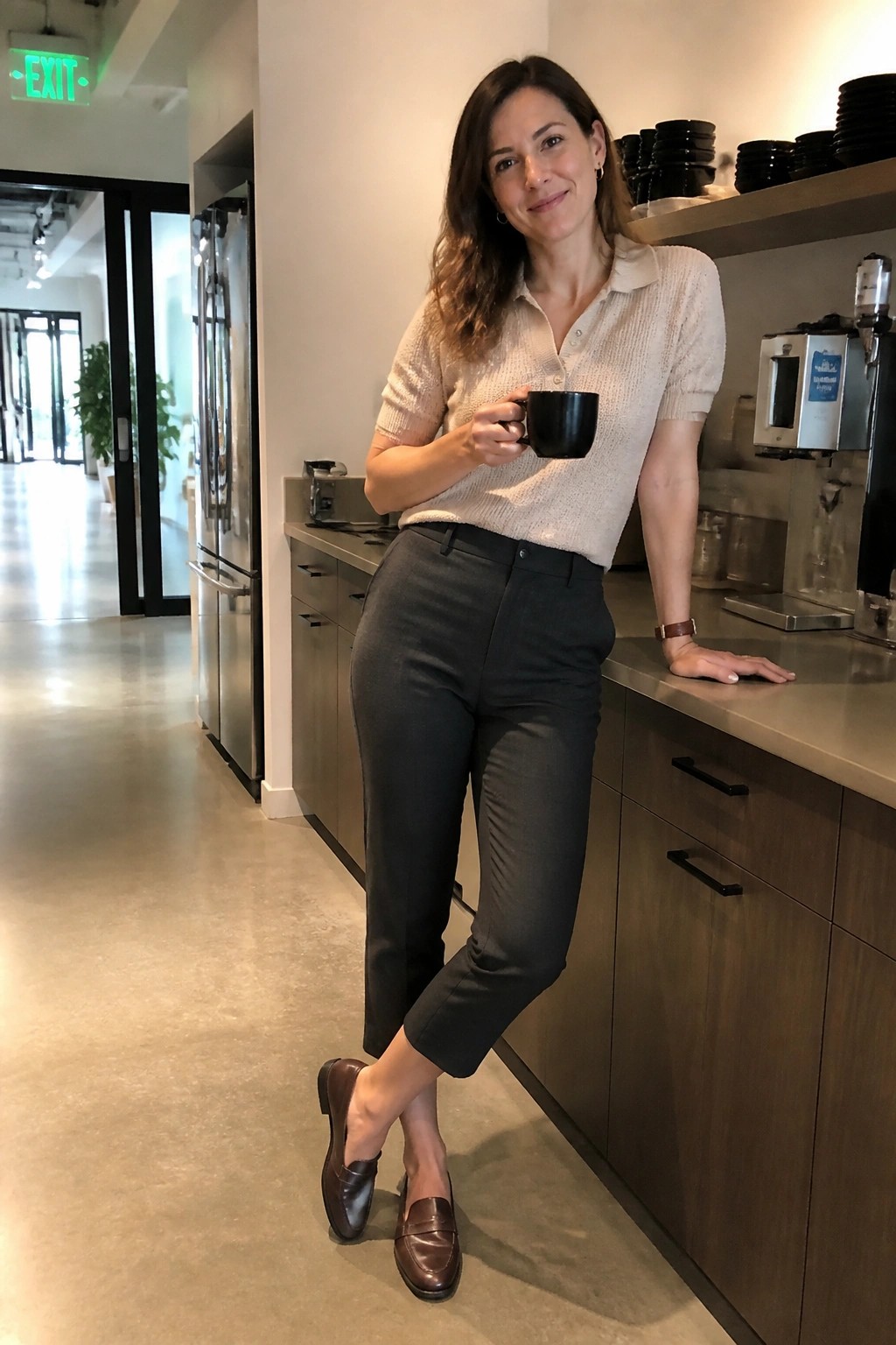 Woman in beige polo sweater and gray pants holding black mug in kitchen.