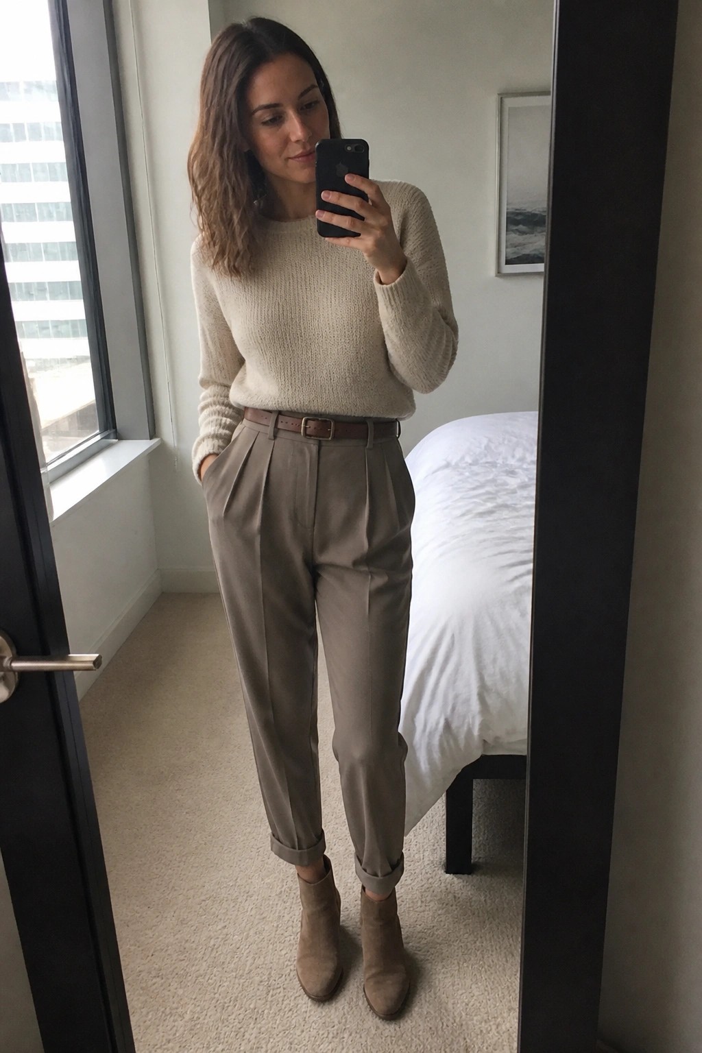 A woman wearing a cream knit sweater tucked into taupe pleated trousers with a brown belt and suede ankle boots while taking a mirror selfie indoors.