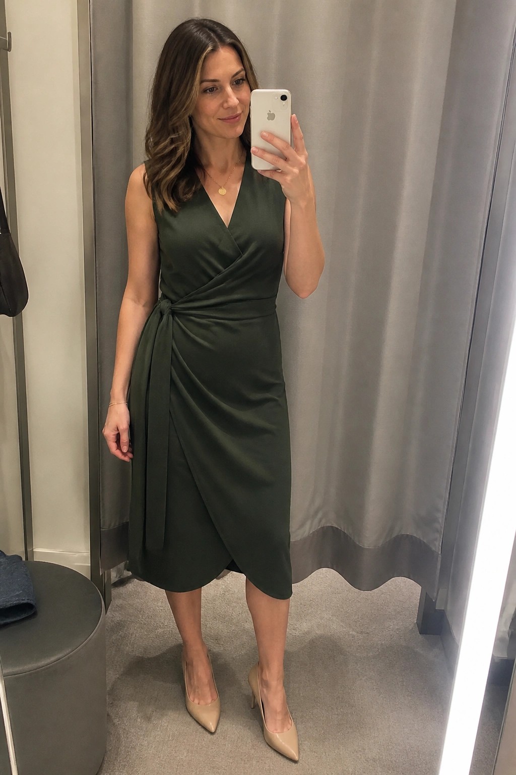 Woman in a sleeveless dark green wrap dress tied at the waist and nude pointed toe heels taking a mirror selfie indoors