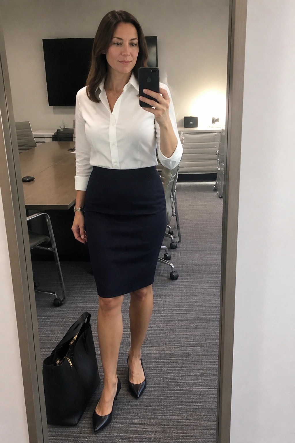 Woman in white shirt and navy pencil skirt taking mirror selfie in office