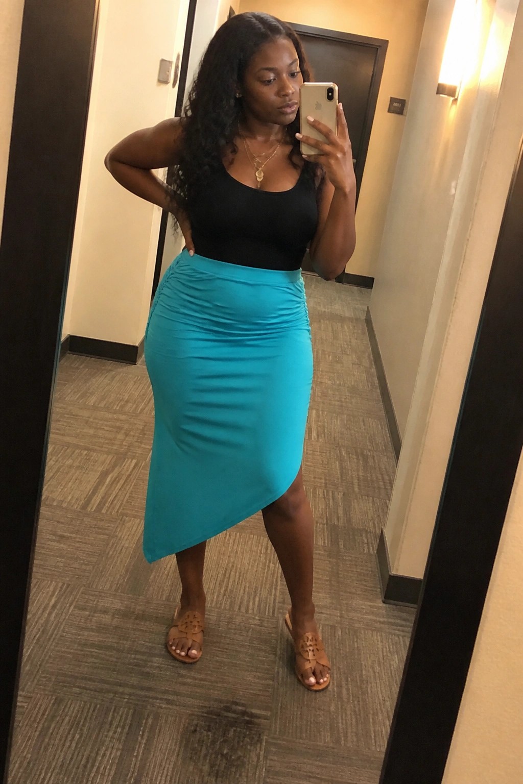 Woman in black top and turquoise ruched skirt taking mirror selfie in hallway