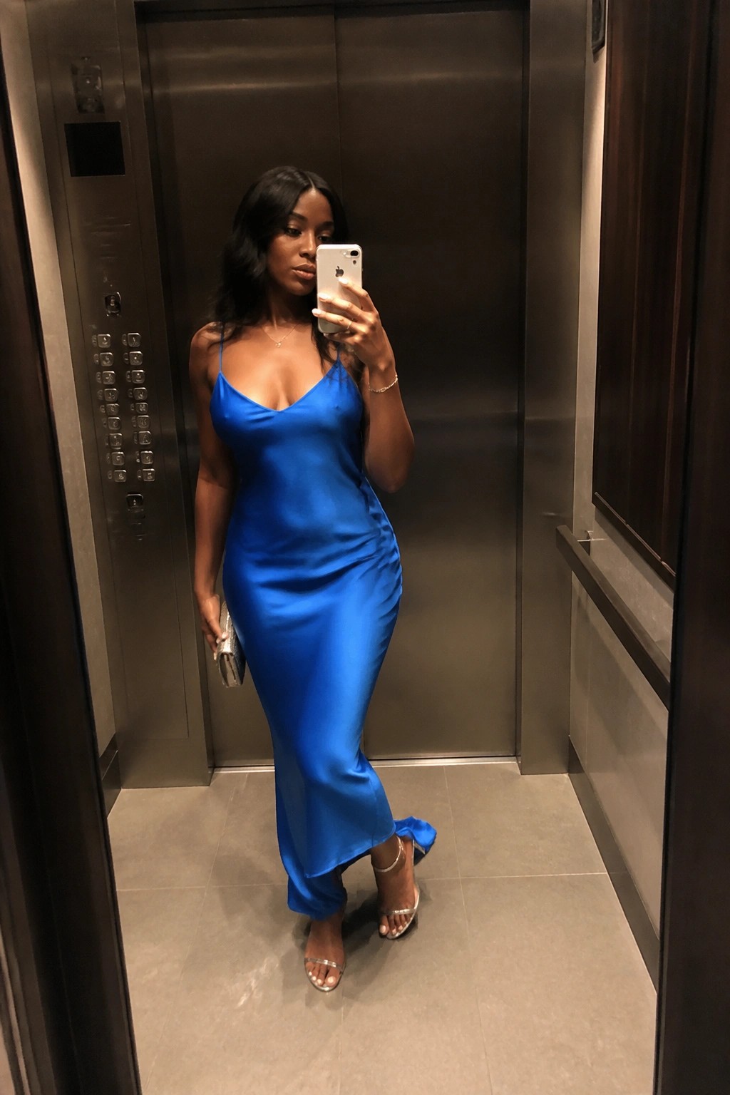 Woman in long blue satin dress taking mirror selfie in elevator