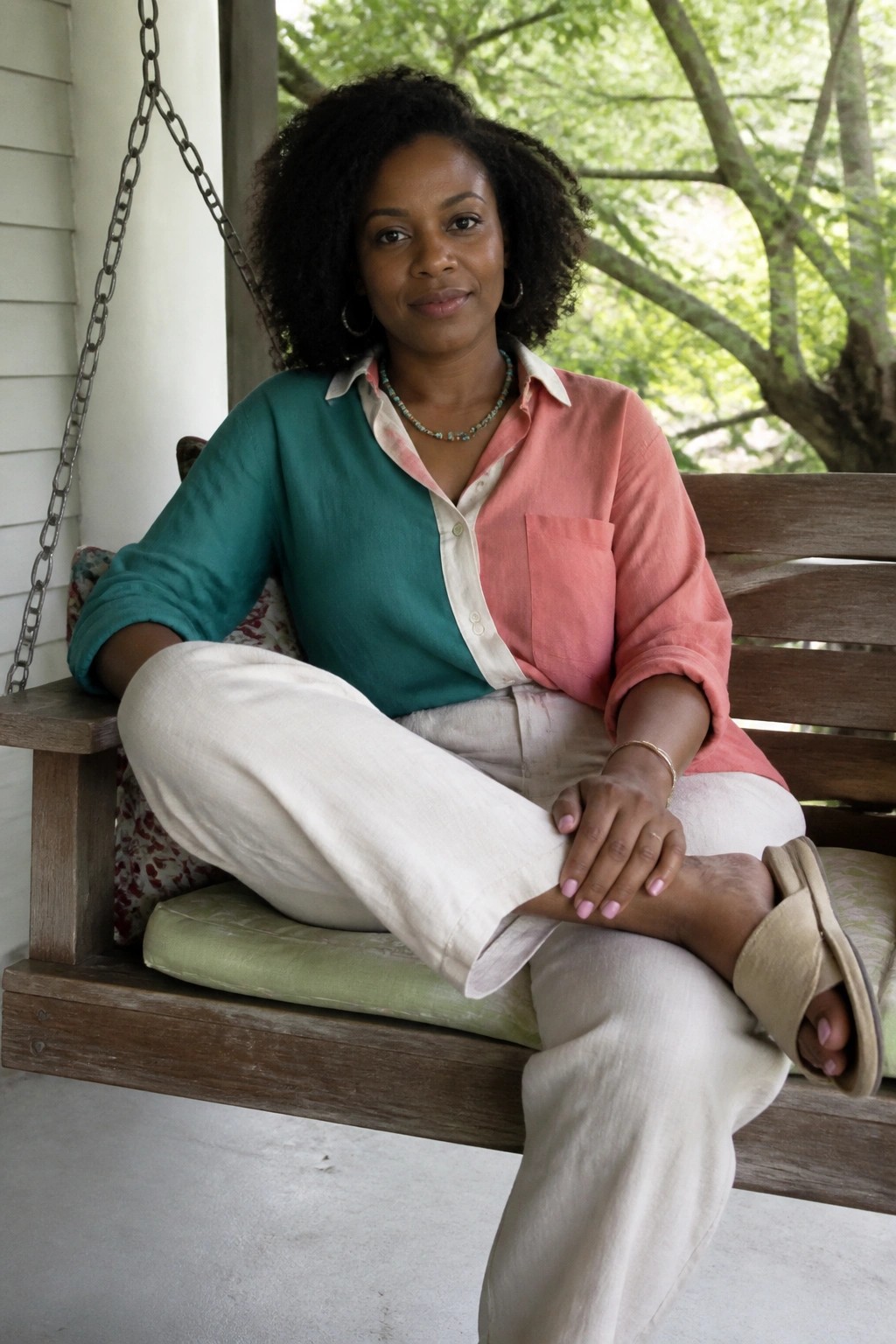 Woman with curly hair in teal-pink shirt seated on wooden porch swing