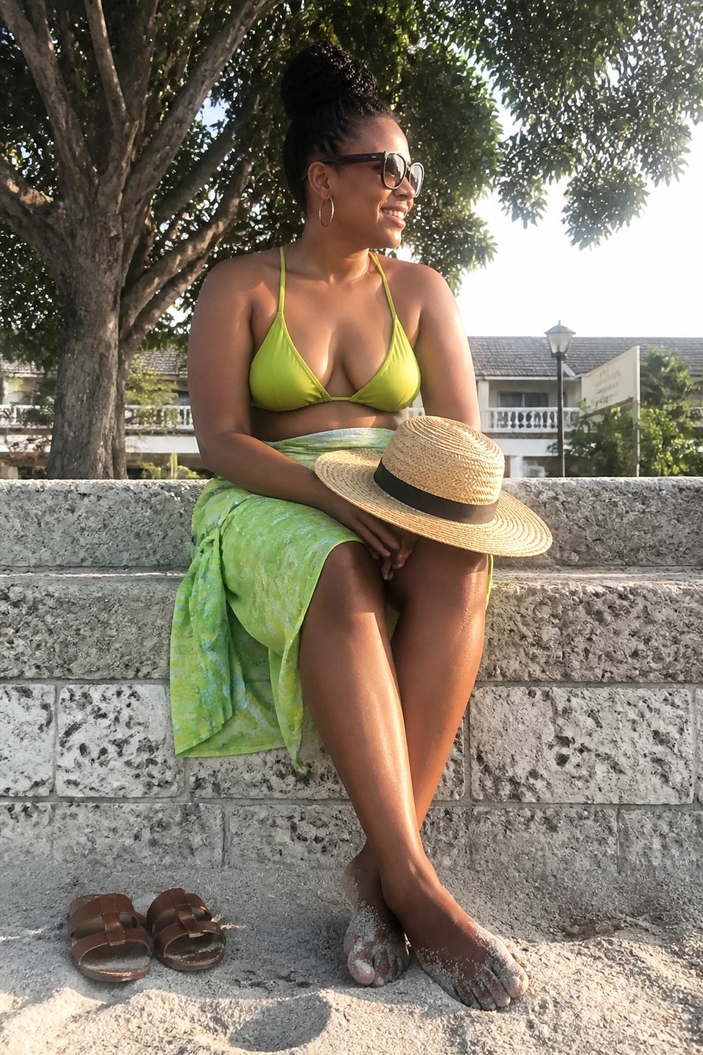 Woman in lime green bikini and skirt sits on stone wall holding straw hat
