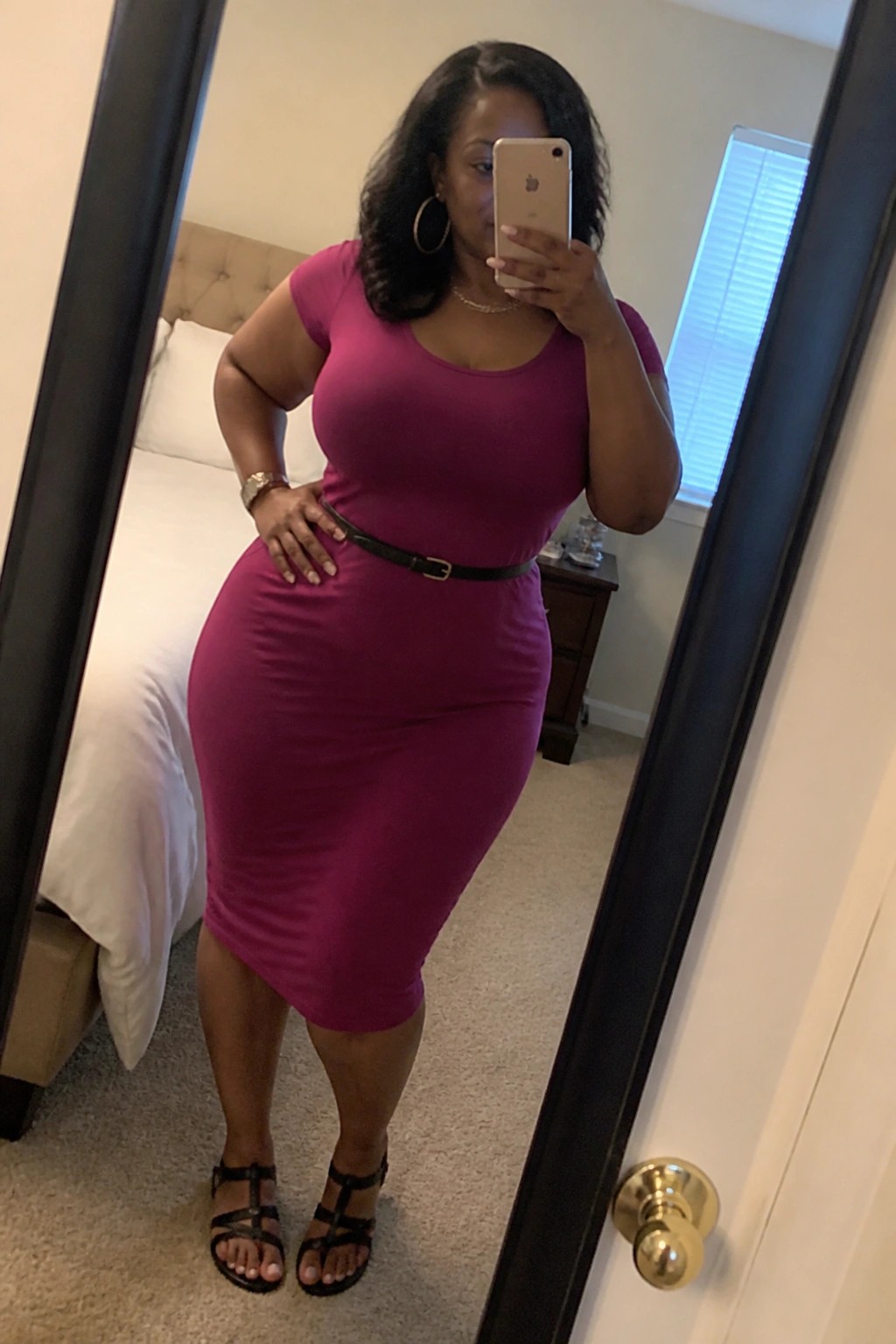 Woman wearing a fitted magenta midi dress with black belt and black strappy sandals taking a mirror selfie in a bedroom setting