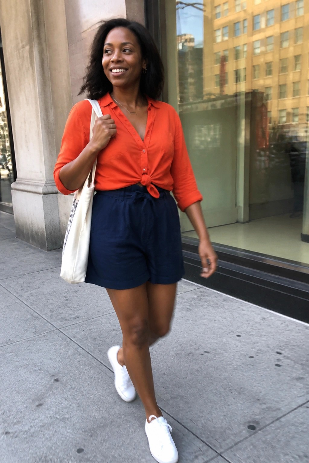 Woman wearing a bright orange button-up shirt tied in a knot at the waist over navy blue shorts with a matching tie waistband, white sneakers, and a white canvas tote bag slung over one shoulder.