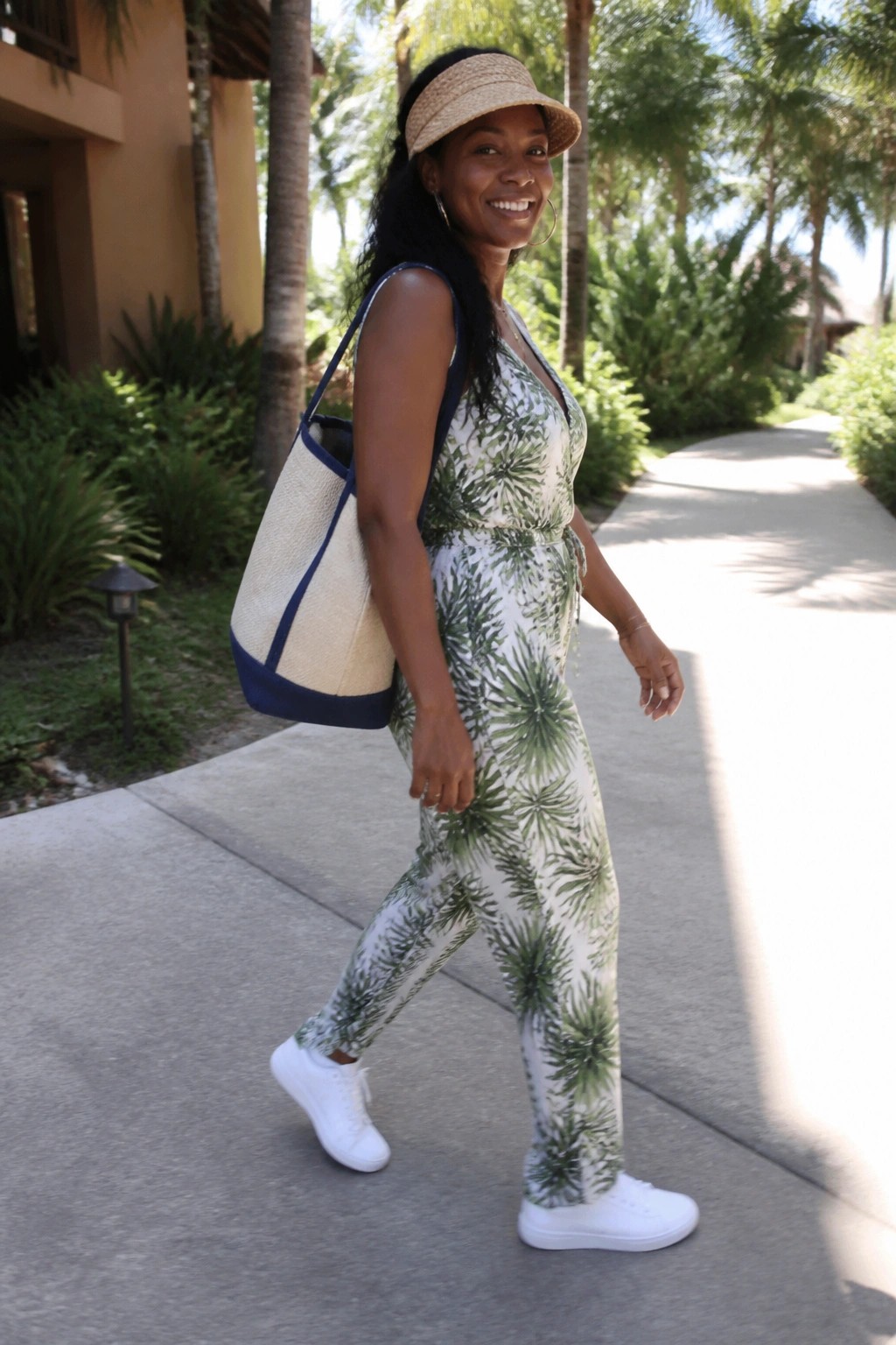 Woman in green palm-print jumpsuit and visor carries woven tote on sunny path