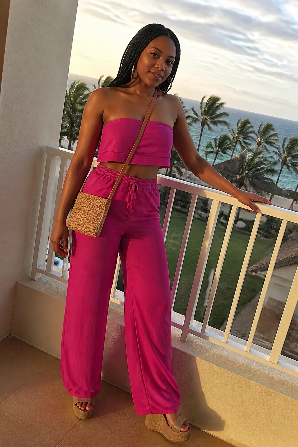 Woman wearing a bright pink strapless crop top and matching wide leg pants with drawstring waist, a woven crossbody bag, and platform wedge sandals on a balcony.