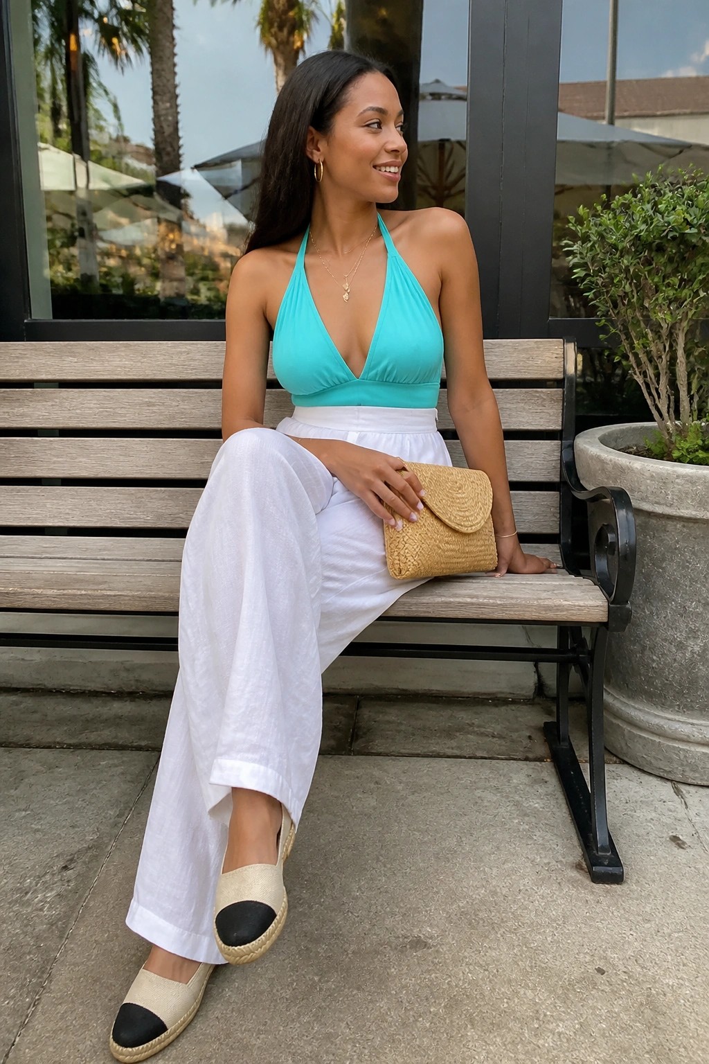 Black woman seated on bench wearing turquoise halter top, white wide leg pants, two-tone espadrilles, and holding a woven straw clutch with gold jewelry.