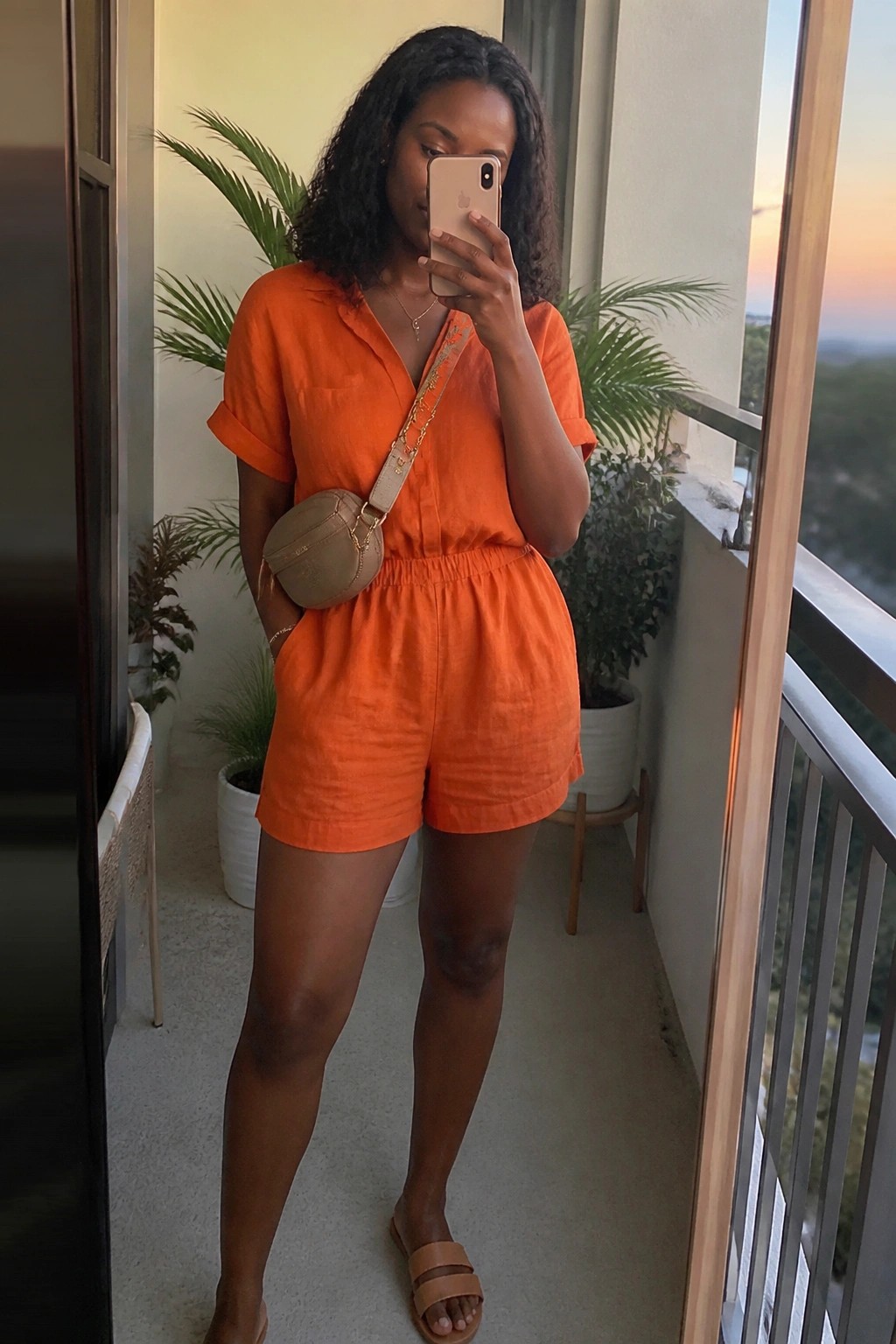 Woman in orange linen romper taking mirror selfie on balcony with crossbody bag
