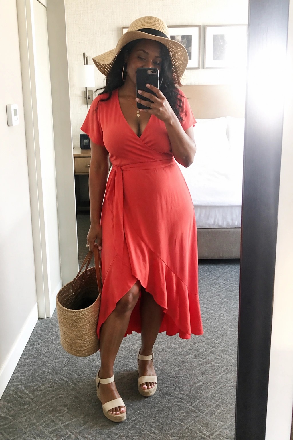 Woman in red wrap dress, straw hat, holding phone and woven bag in mirror