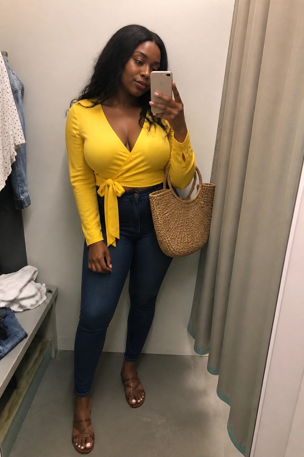 Woman in mirror selfie wearing bright yellow long sleeve wrap top tied at waist with dark blue jeans brown strappy sandals and carrying a large woven straw tote bag.
