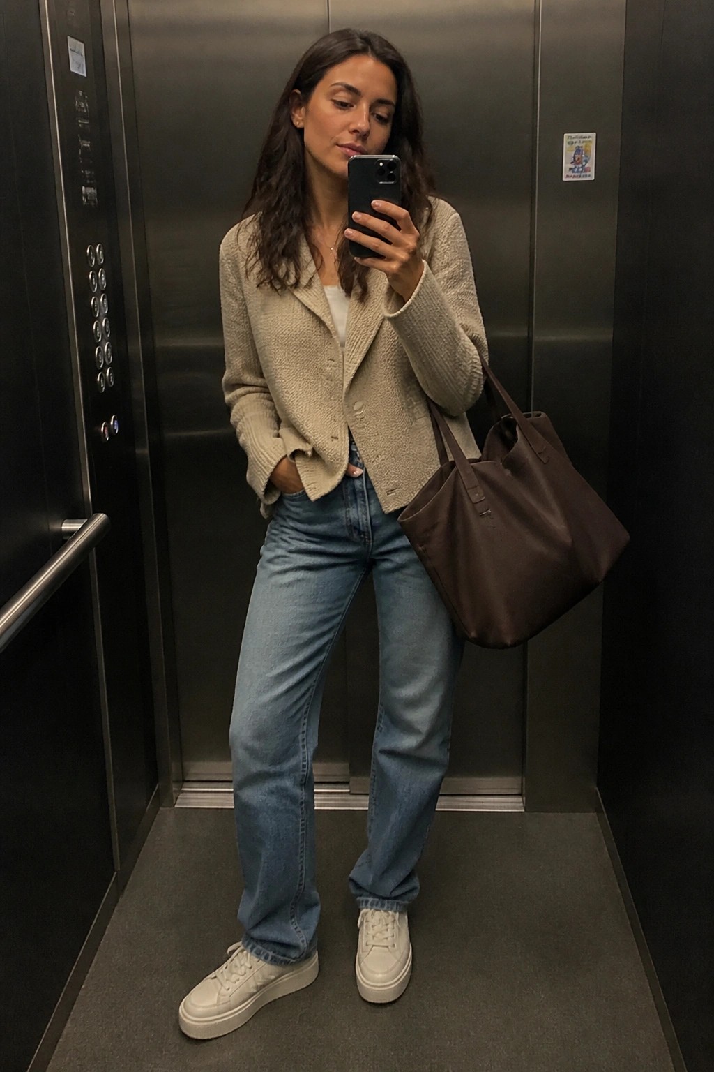 Woman in beige cardigan and jeans taking selfie in elevator with brown tote bag