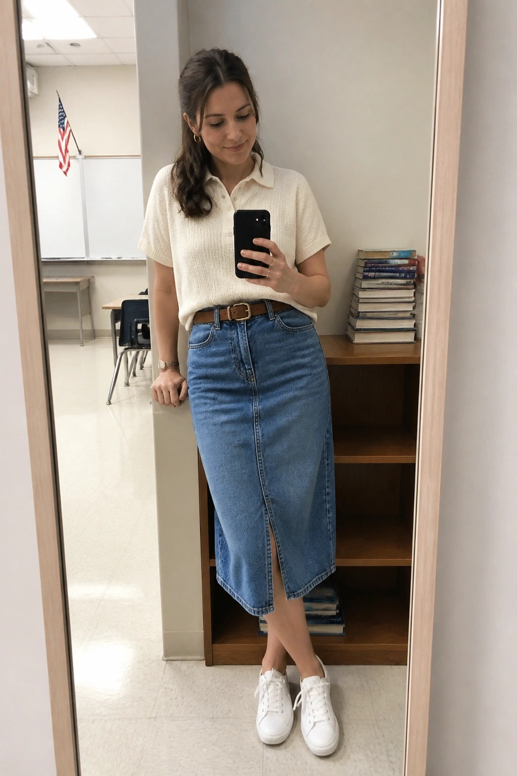 Woman wearing cream short sleeve knit polo sweater tucked into blue denim midi skirt with brown belt and white sneakers standing in classroom setting holding phone.