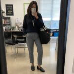 Woman wearing a navy blue sweater tucked into cropped gray trousers, paired with black loafers featuring gold hardware and carrying a black tote bag with a lanyard around her neck.