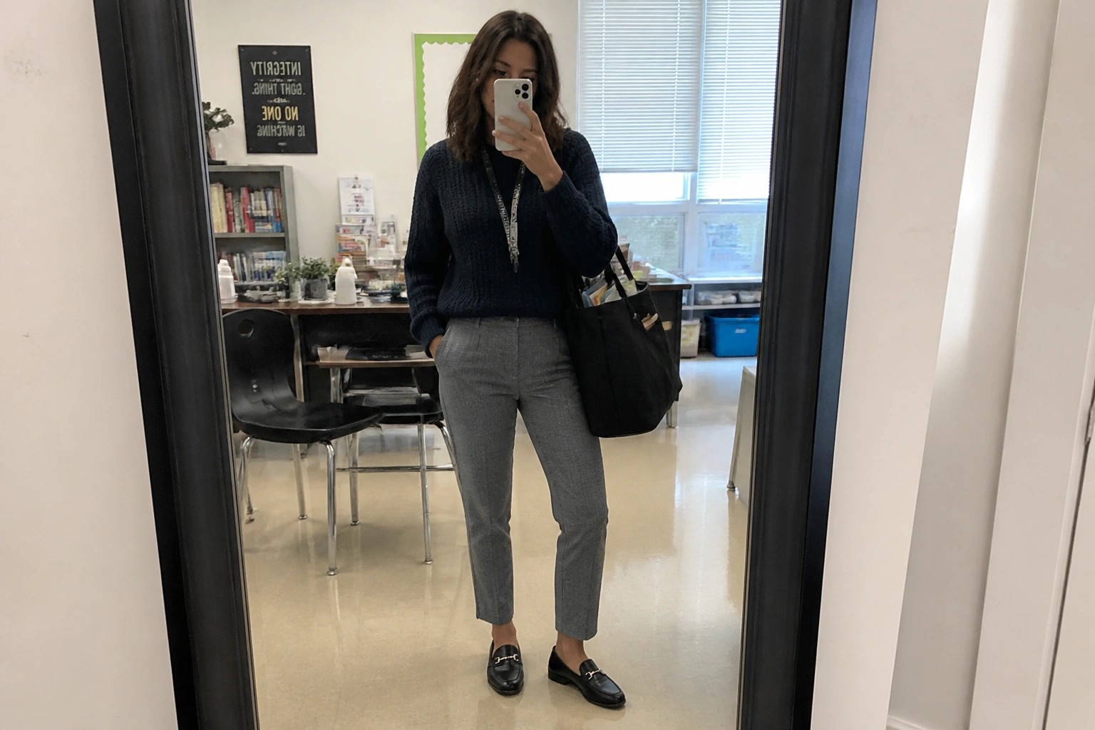 Woman wearing a navy blue sweater tucked into cropped gray trousers, paired with black loafers featuring gold hardware and carrying a black tote bag with a lanyard around her neck.