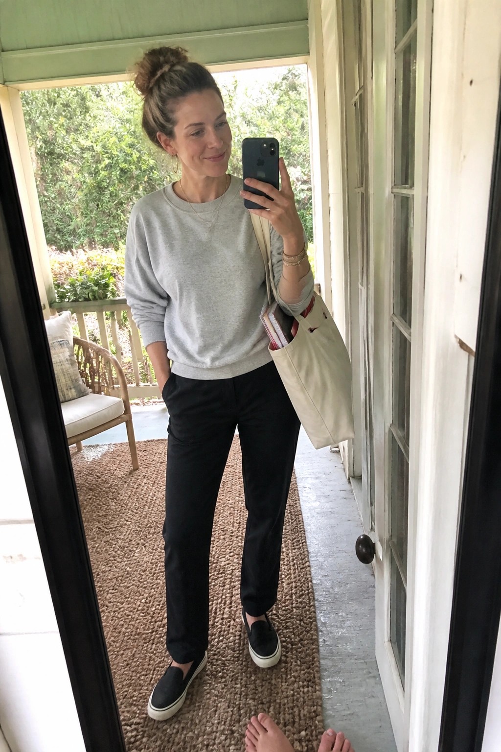 Woman in gray sweatshirt and black pants taking mirror selfie with beige tote bag