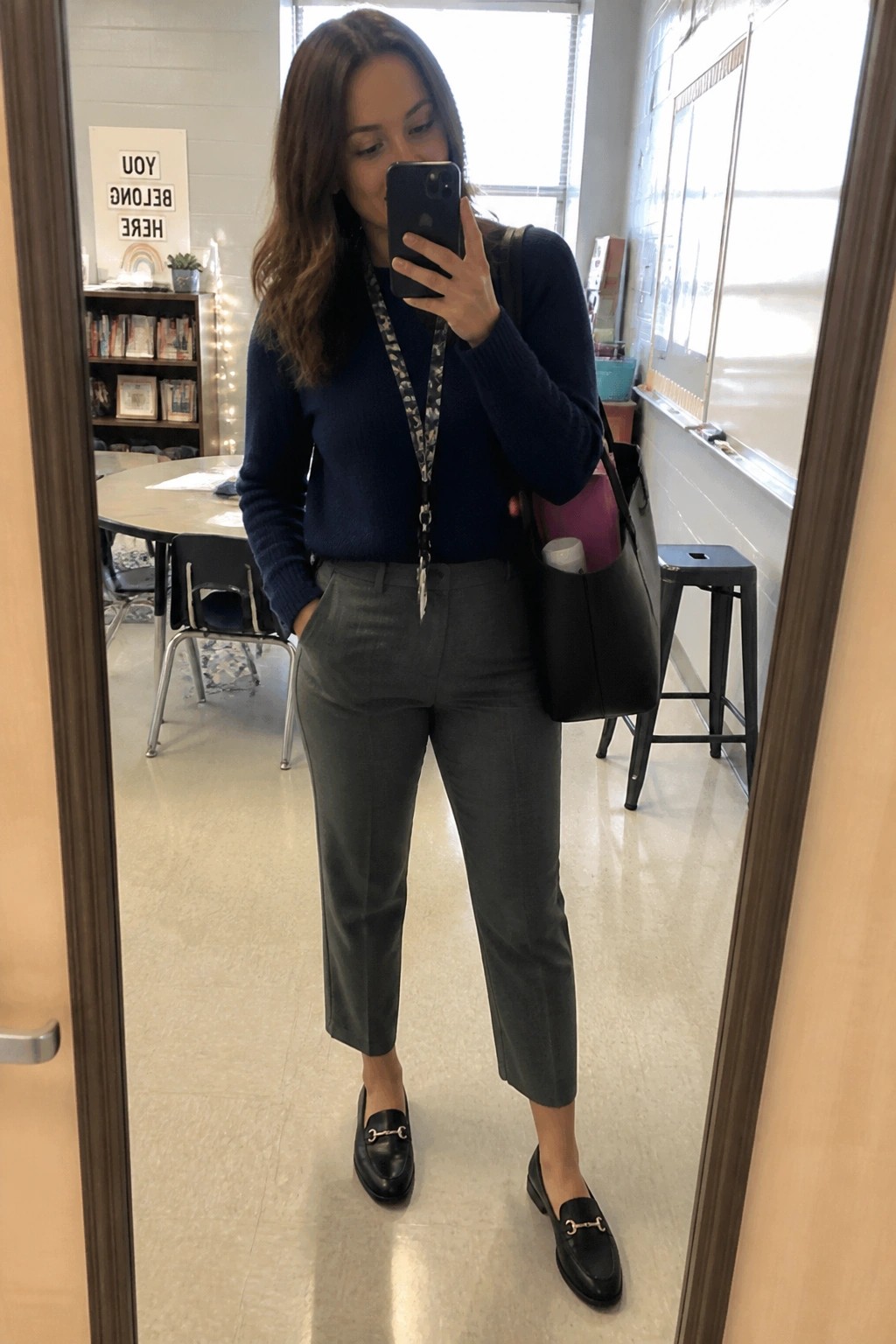 Woman wearing a navy blue sweater tucked into cropped gray trousers, paired with black loafers featuring gold hardware and carrying a black tote bag with a lanyard around her neck.