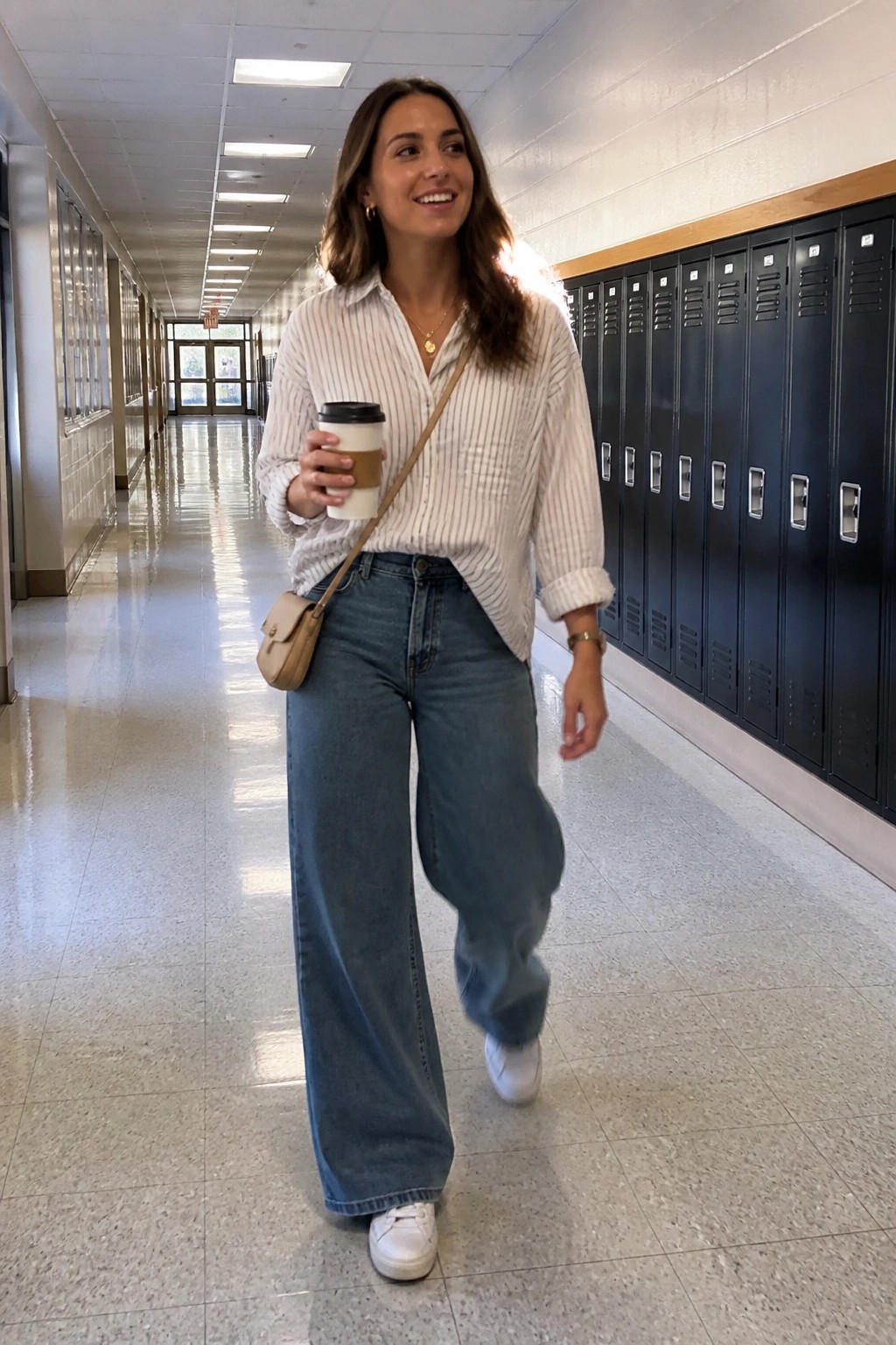 Woman in striped shirt and wide-leg jeans holding coffee in locker hallway