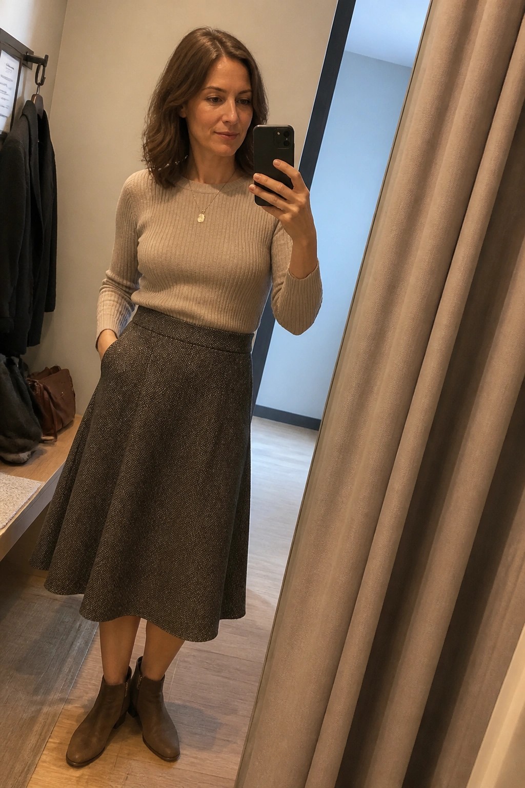 Woman in a beige ribbed knit sweater tucked into a dark speckled A-line midi skirt paired with brown ankle boots and a thin gold necklace while taking a mirror selfie.