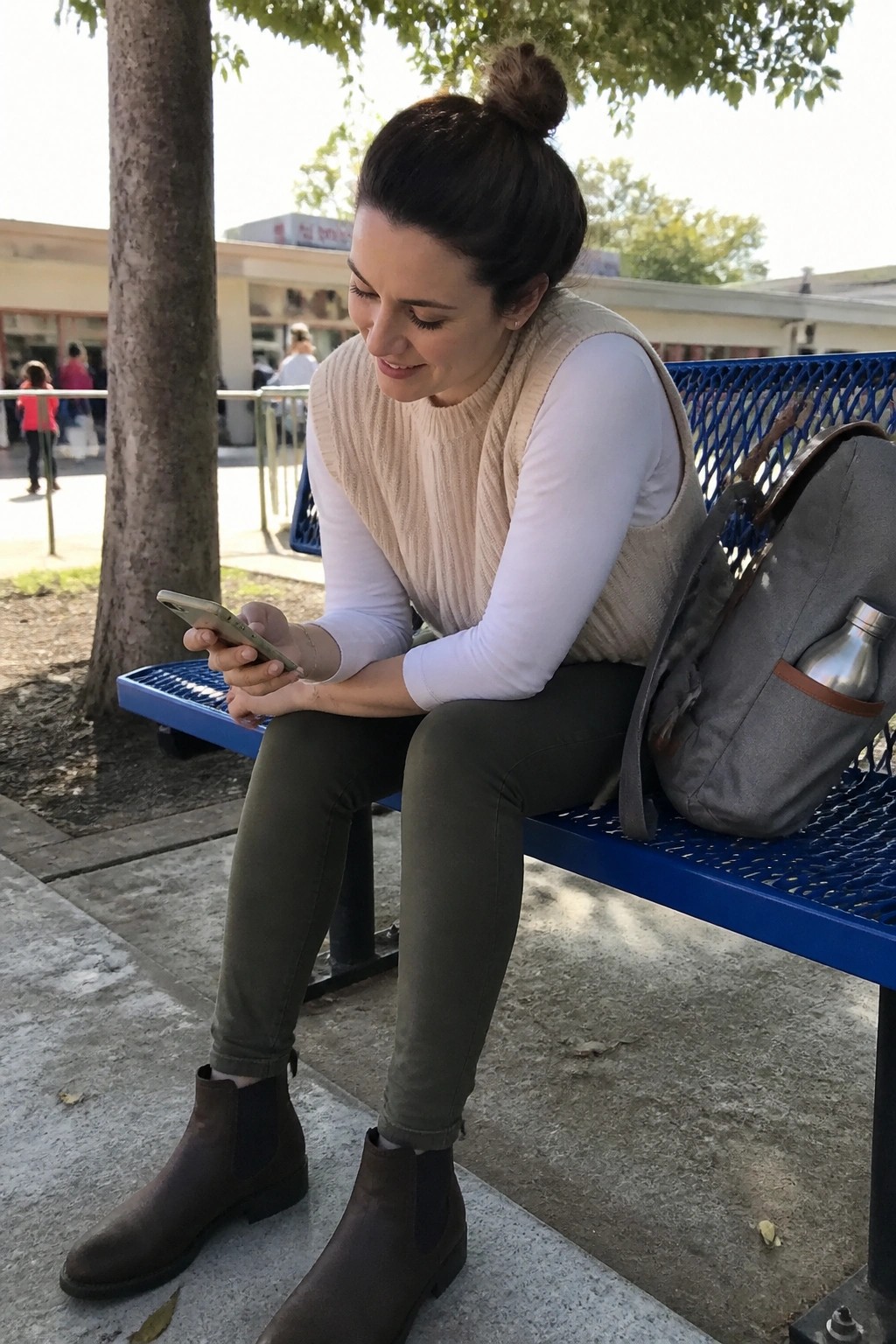 Woman in beige vest sits on blue bench viewing smartphone beside backpack