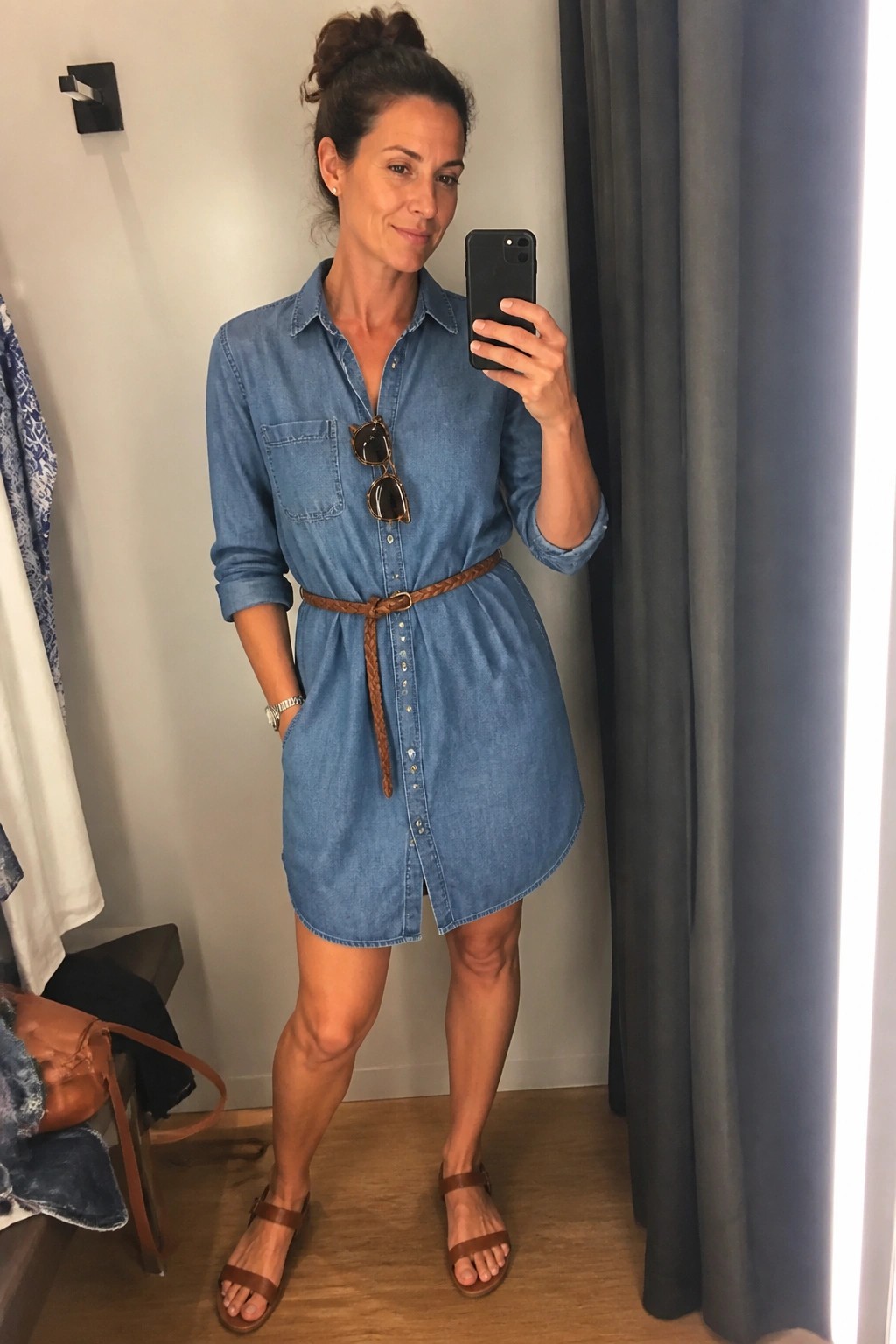 Woman wearing a blue denim shirt dress belted at the waist with a brown braided belt, brown sandals, and sunglasses hanging from the front, holding a phone for a mirror selfie.