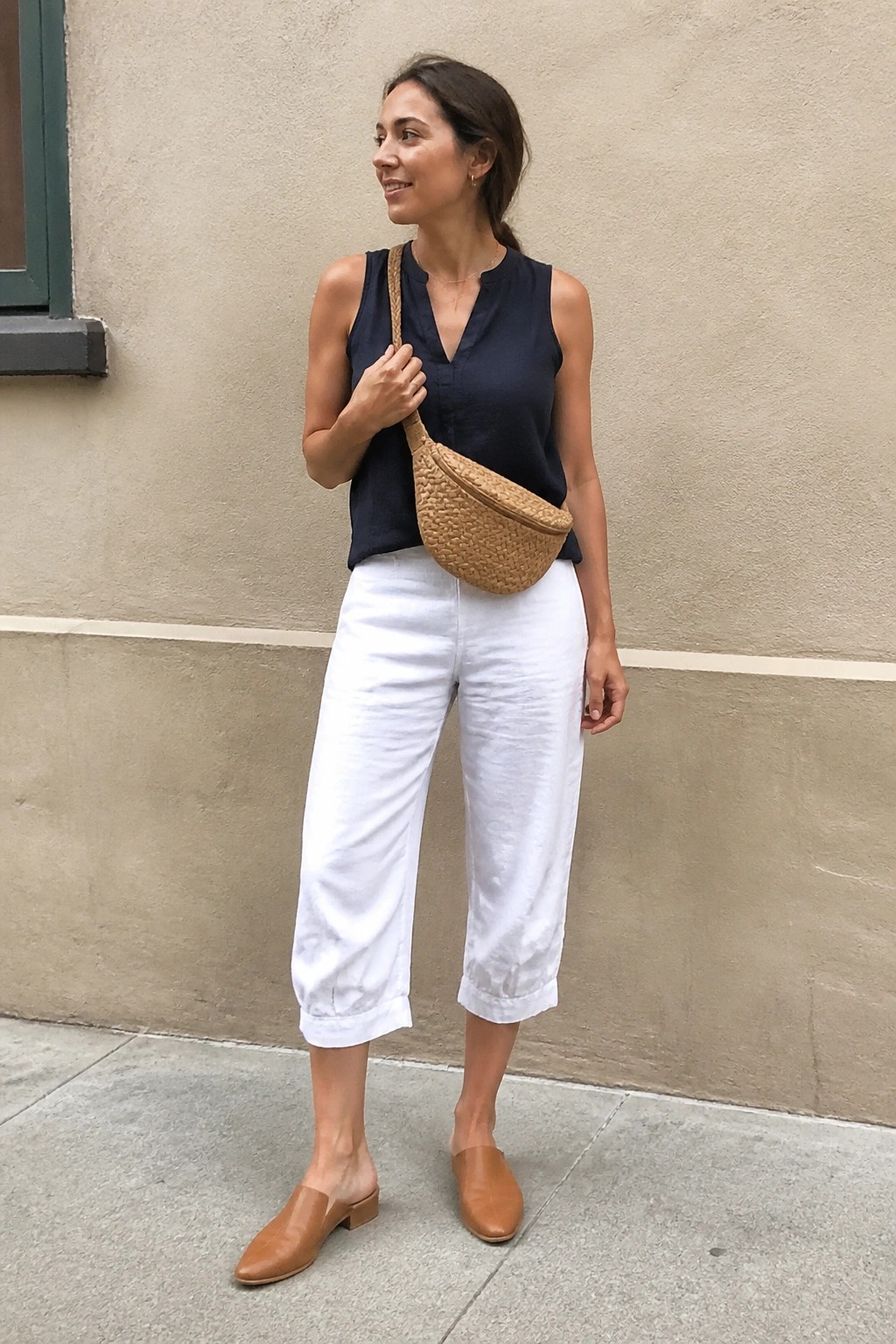 Woman in navy top and white cropped pants with woven fanny pack and brown mules