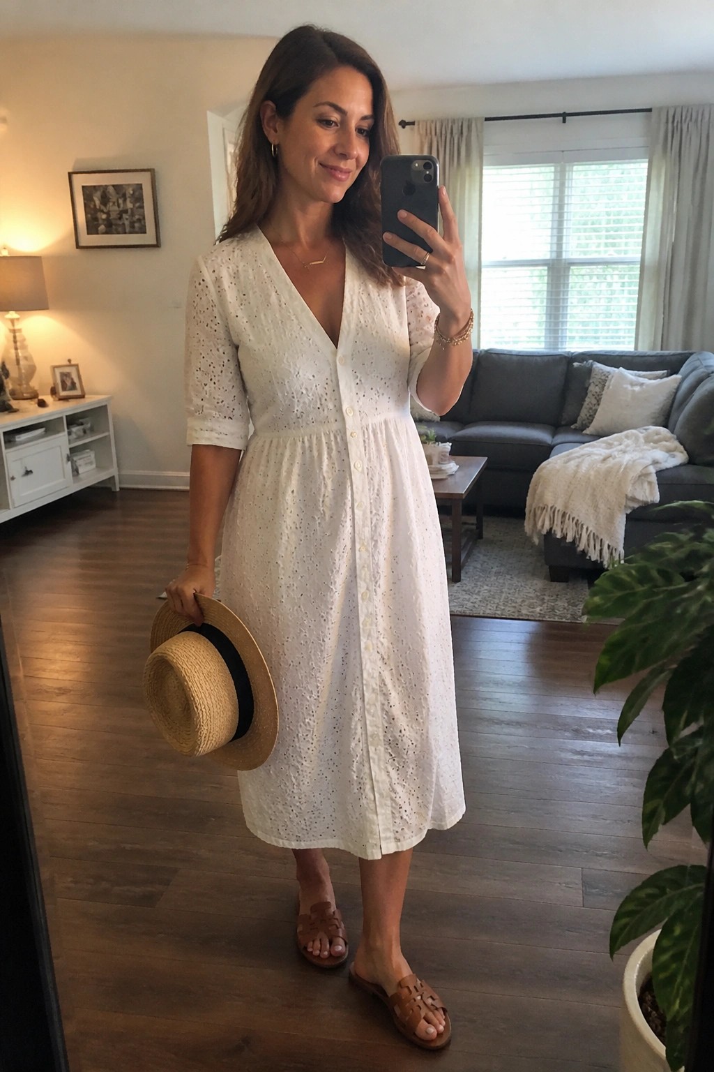 Woman in white eyelet midi dress holds straw hat during mirror selfie in living room
