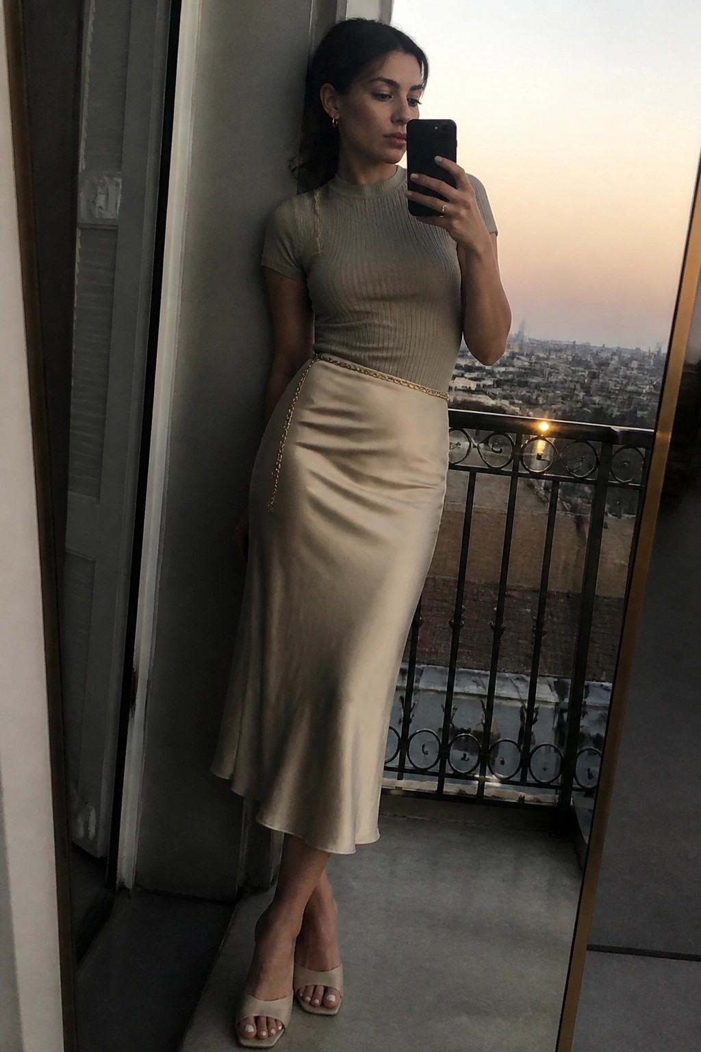Woman wearing a beige ribbed short-sleeve top and beige satin midi skirt with gold chain belt posing in a mirror selfie on a balcony.