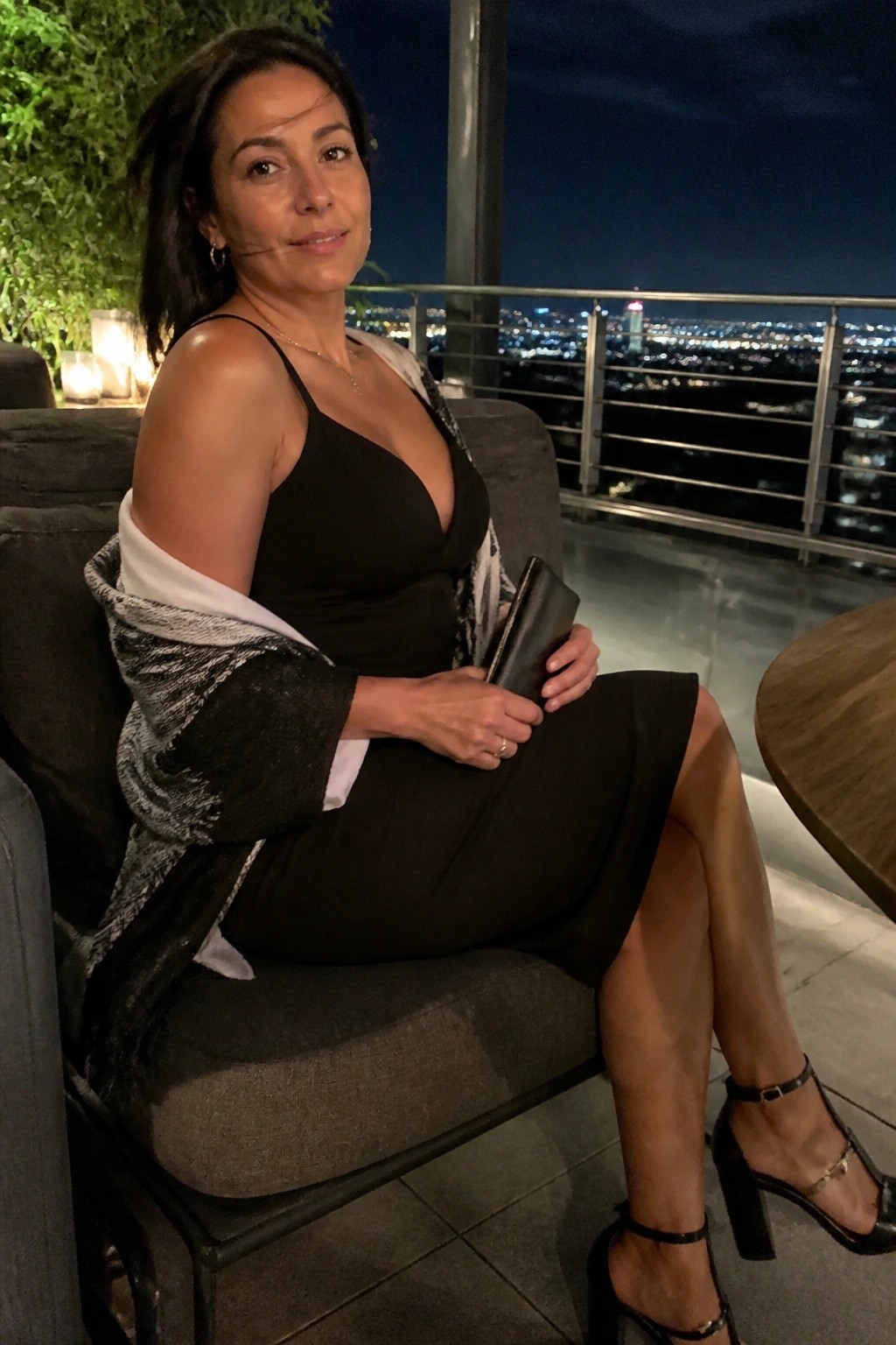 Woman in black dress with patterned shawl on nighttime balcony overlooking city lights.