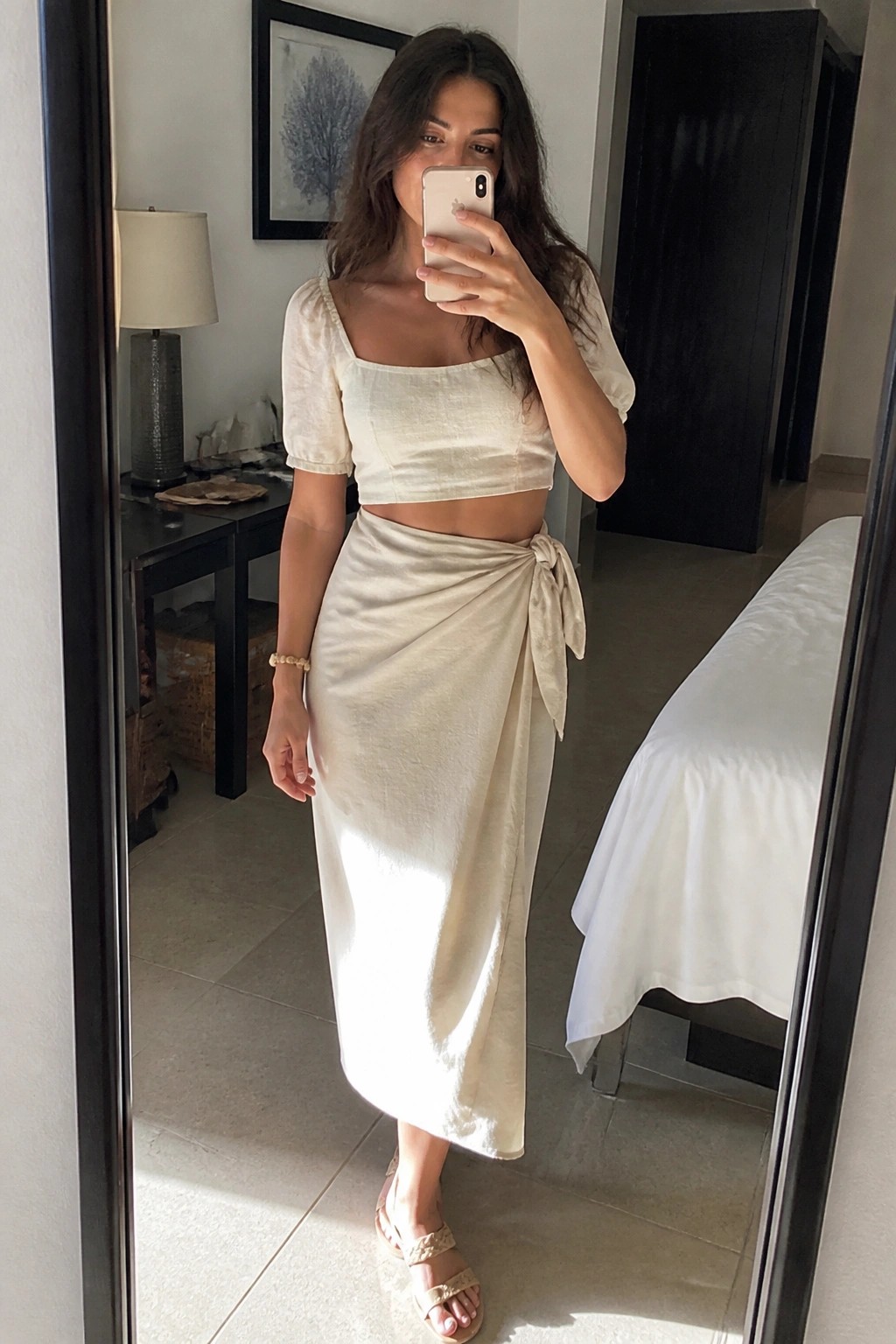 Woman in cream linen crop top and wrap skirt taking bedroom mirror selfie