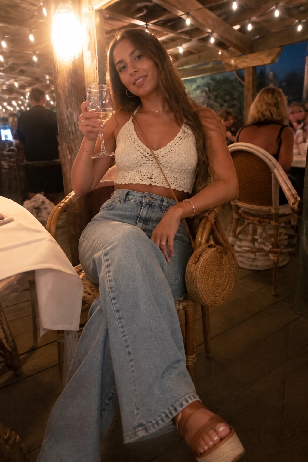 Woman in white crochet top and jeans holding wine glass at outdoor restaurant.