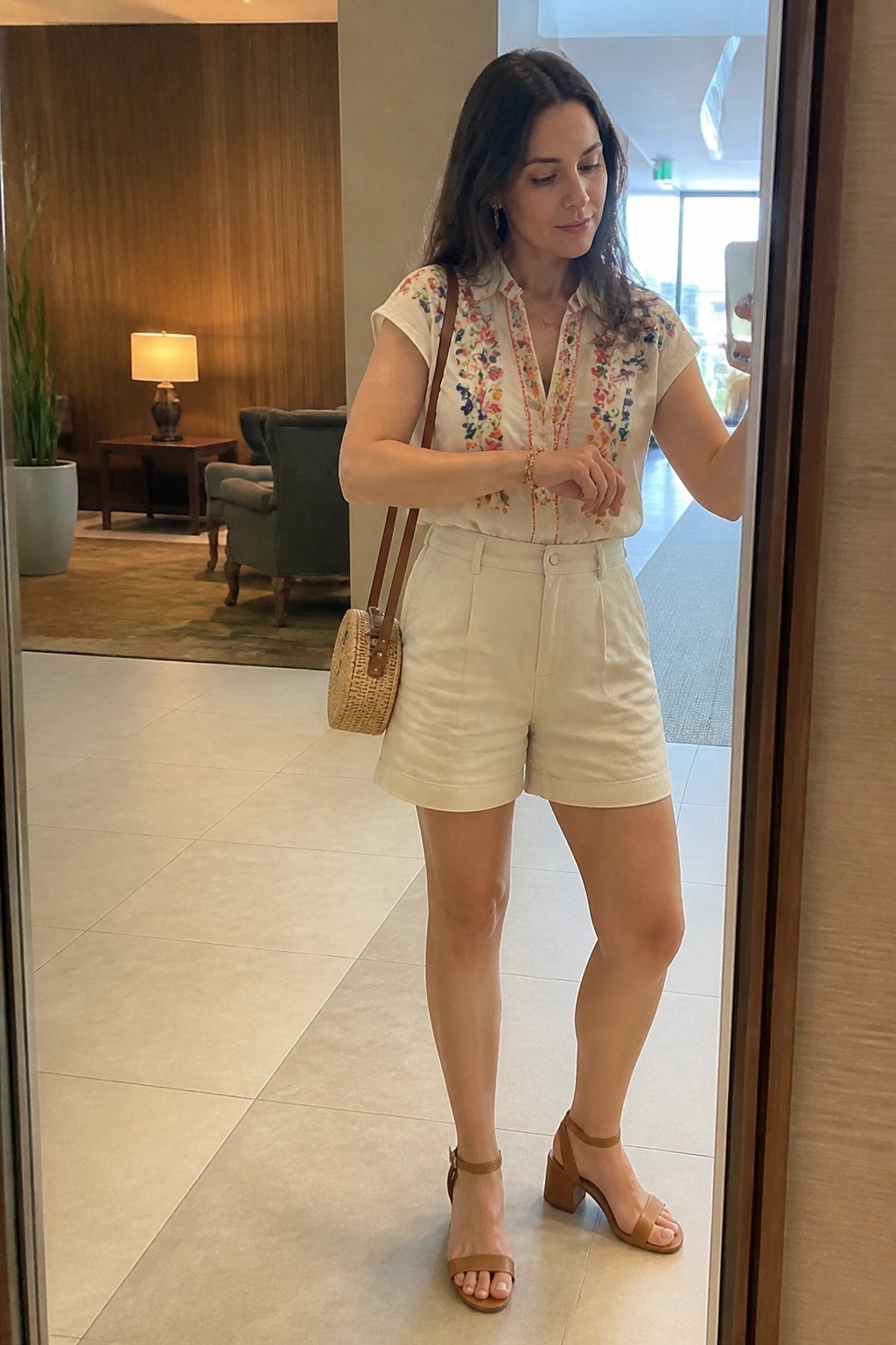 Woman in cream floral-embroidered top and shorts with woven bag taking mirror selfie