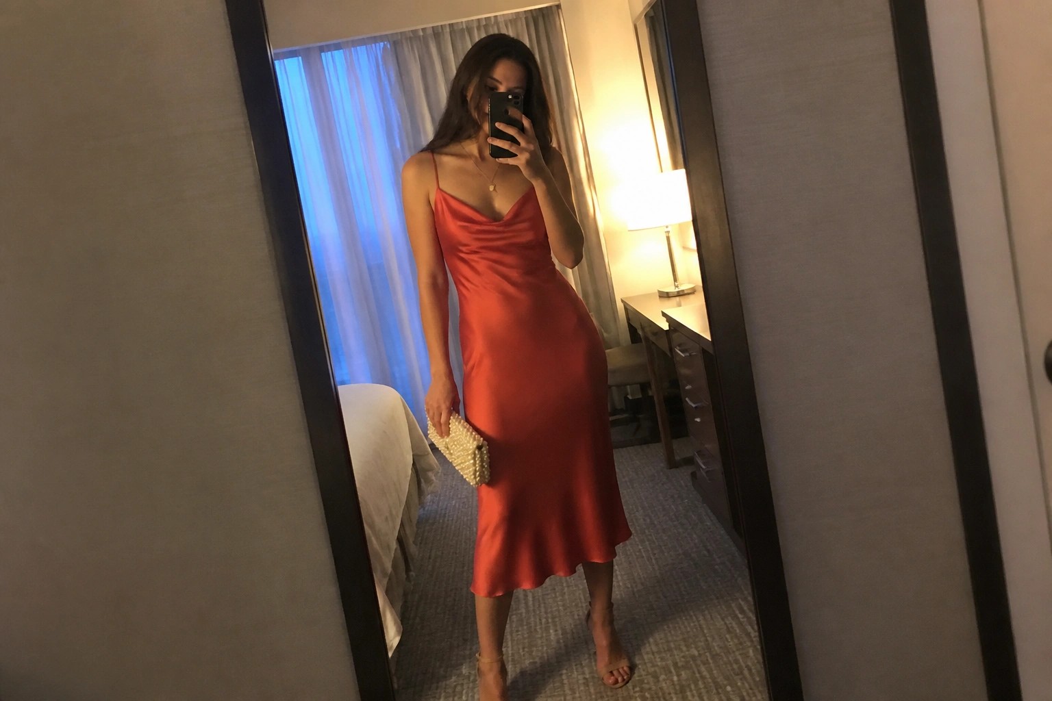 Woman wearing a coral satin midi slip dress with thin straps, cowl neck, holding a gold beaded clutch, paired with nude strappy heels and a delicate gold necklace in a mirror selfie.