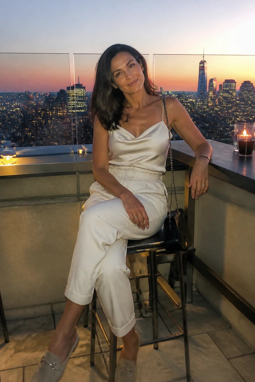 Woman in white satin top seated on rooftop stool overlooking dusk city skyline