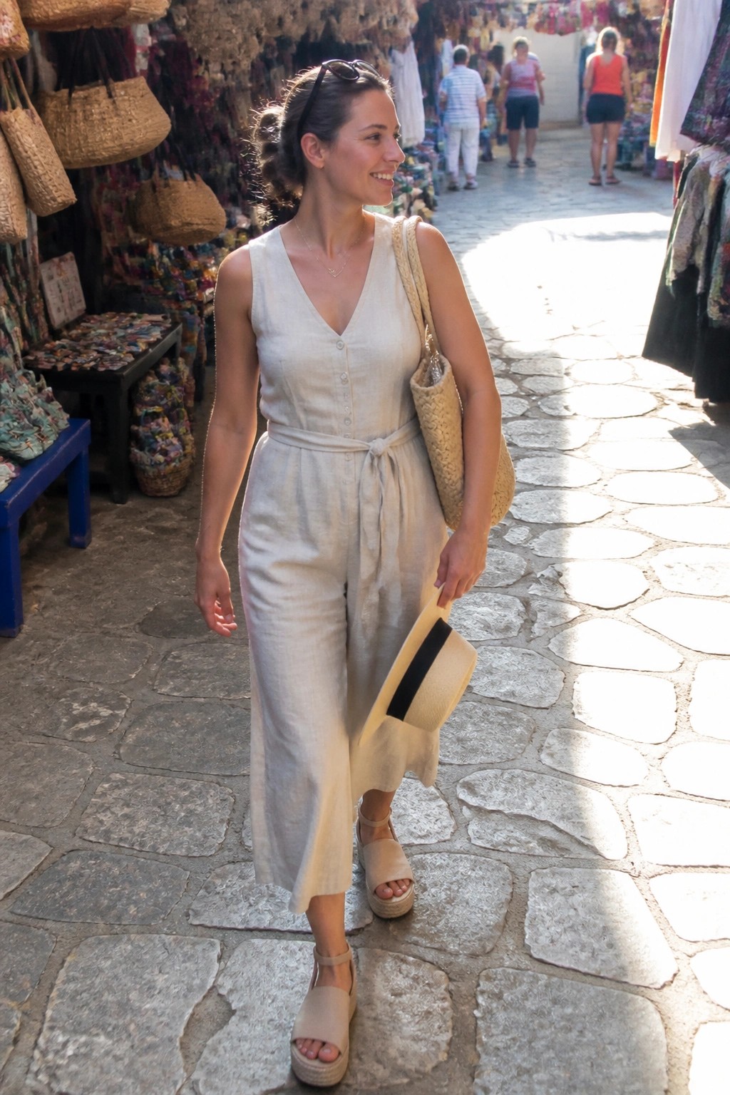 Woman in beige linen jumpsuit holding hat on sunny cobblestone market street