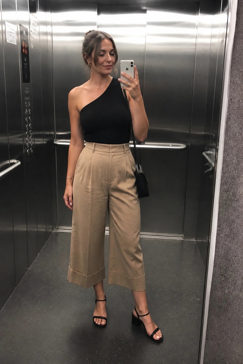 Woman wearing a black one shoulder top with beige wide leg cropped pants and black block heel sandals, holding a phone for a mirror selfie in an elevator.