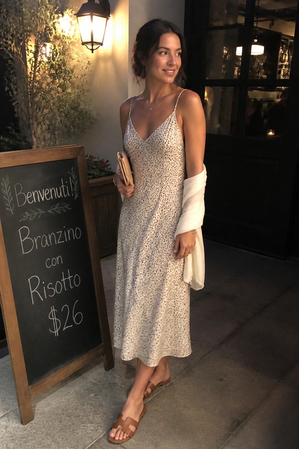 Woman in white speckled dress with sweater beside chalkboard menu outside restaurant at night