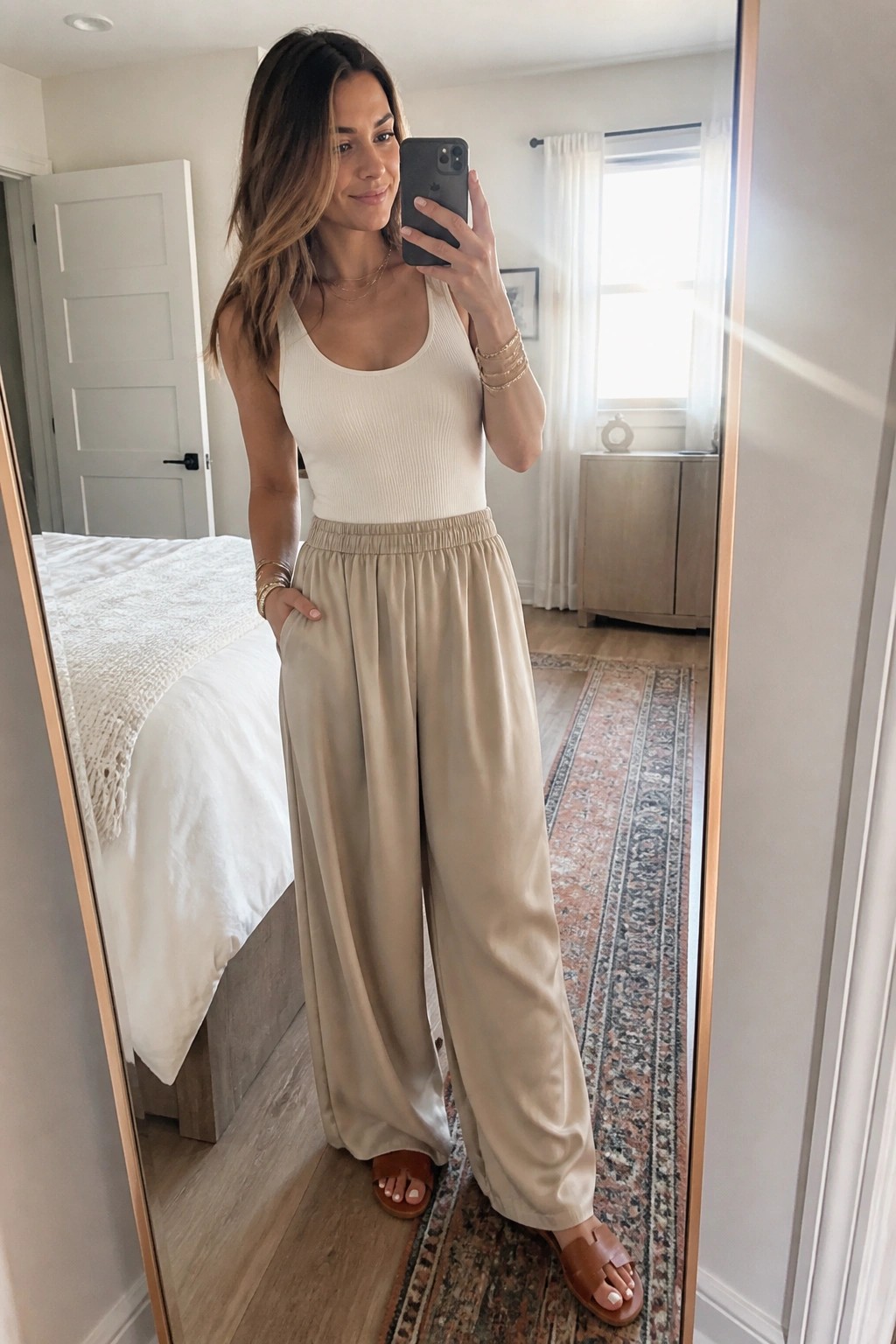 Woman in white ribbed tank and beige wide-leg pants taking bedroom mirror selfie
