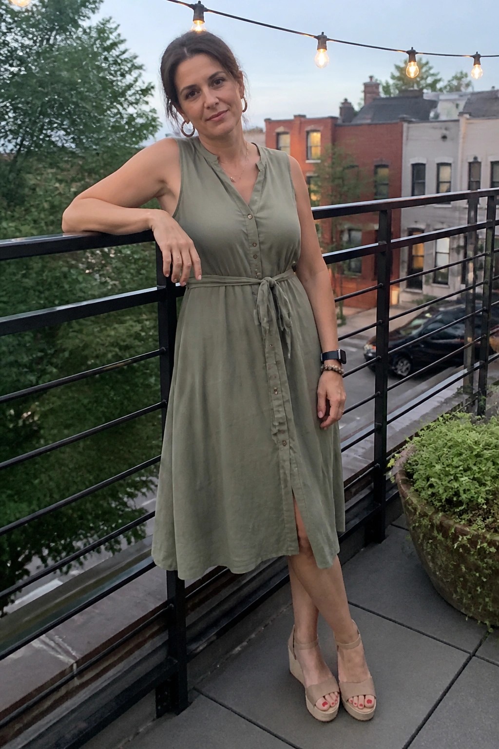 Woman in olive green sleeveless midi dress leaning on urban balcony railing at dusk