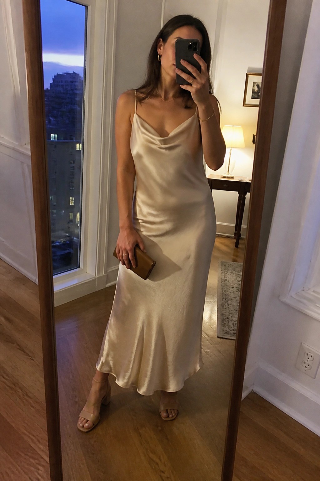 Woman in cream satin slip dress taking mirror selfie with phone and clutch