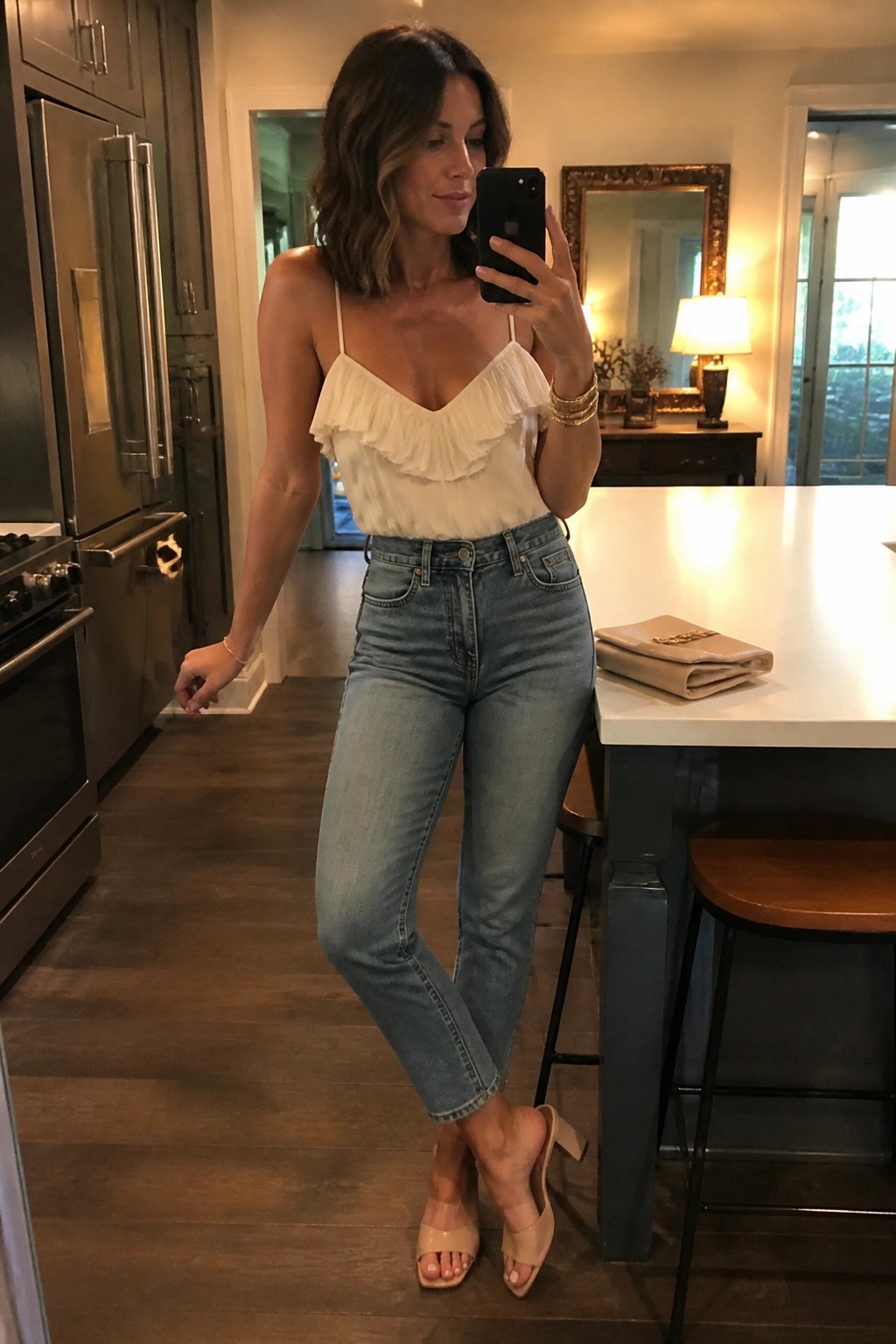 Woman in white ruffled camisole, light blue straight leg jeans and beige heeled mules posing in a kitchen setting