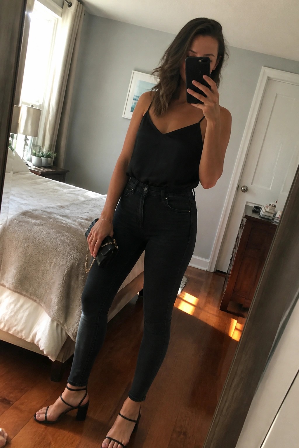 Woman in black camisole and jeans taking mirror selfie in bedroom